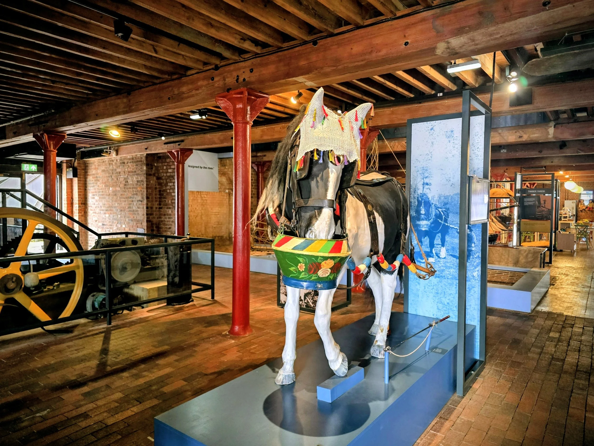 Decorated horse sculpture display inside a museum or gallery with wooden beams, brick walls, and informational panels.