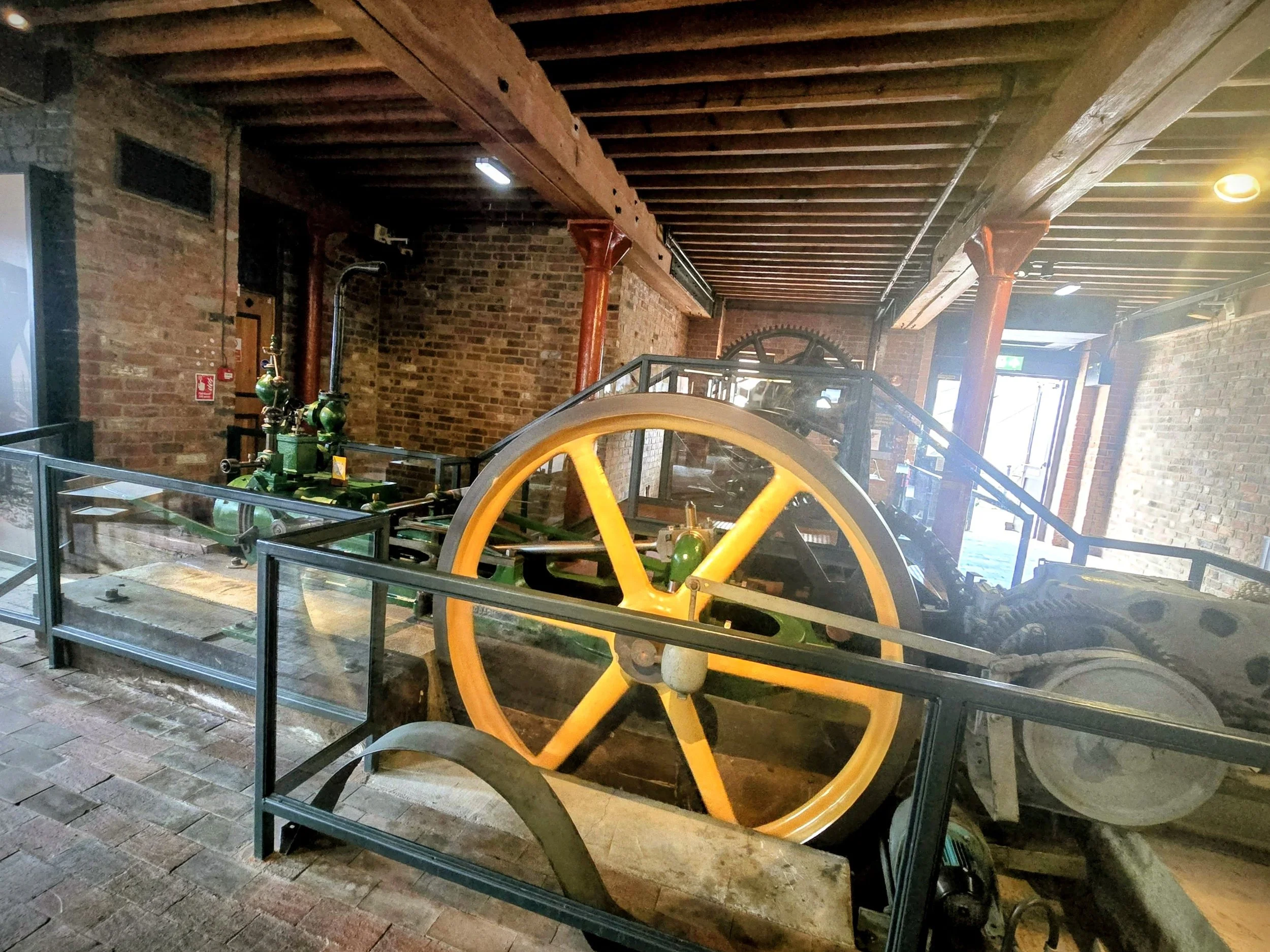 Historical industrial machinery with large yellow wheel, green and black components, enclosed by glass and metal barriers inside a brick building with wooden beams.