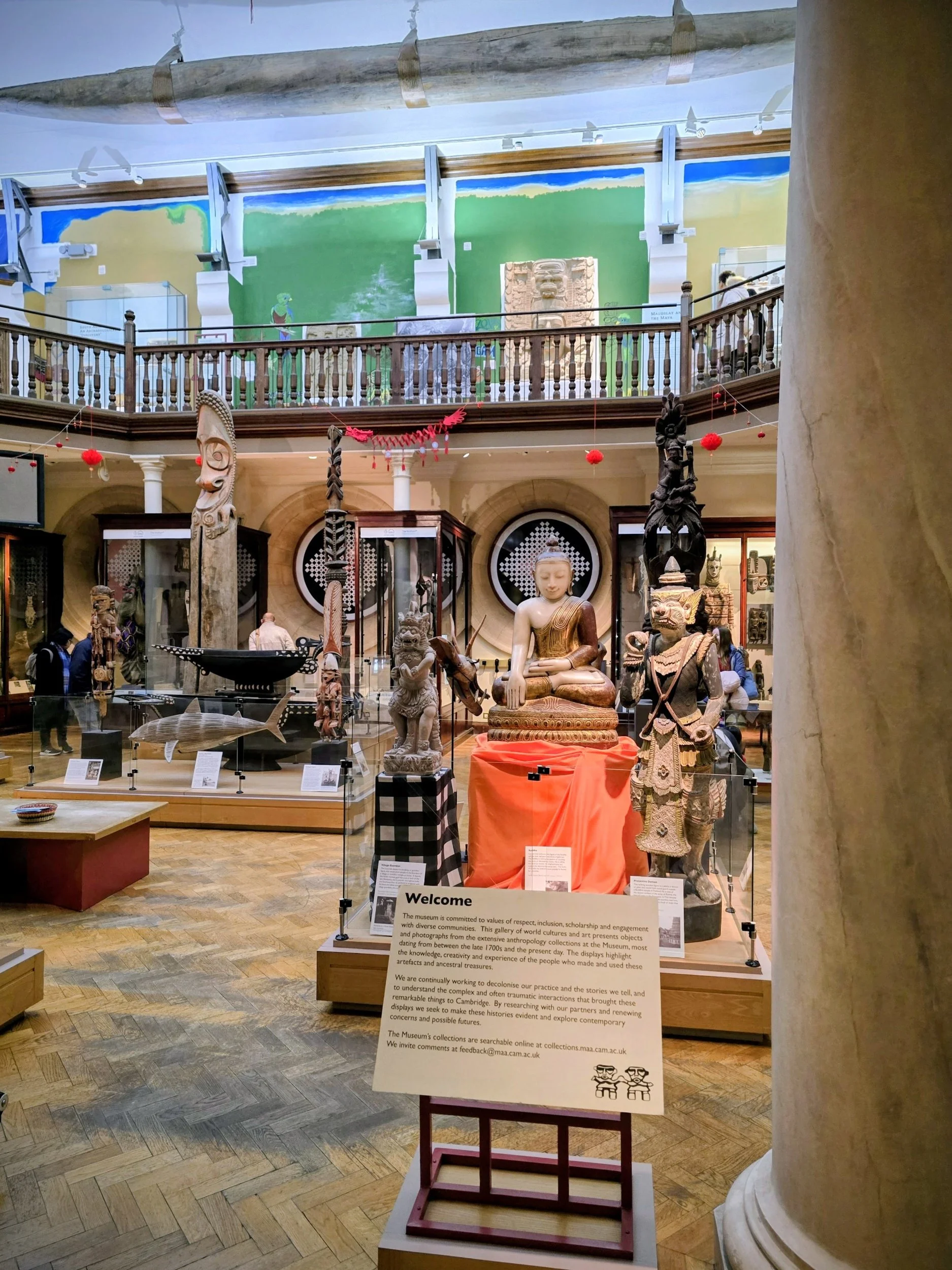 Interior of a museum exhibit hall with various sculptures and artifacts, including a large seated figure, a fish model, and statues of warriors, under a balcony with artwork on the upper wall.