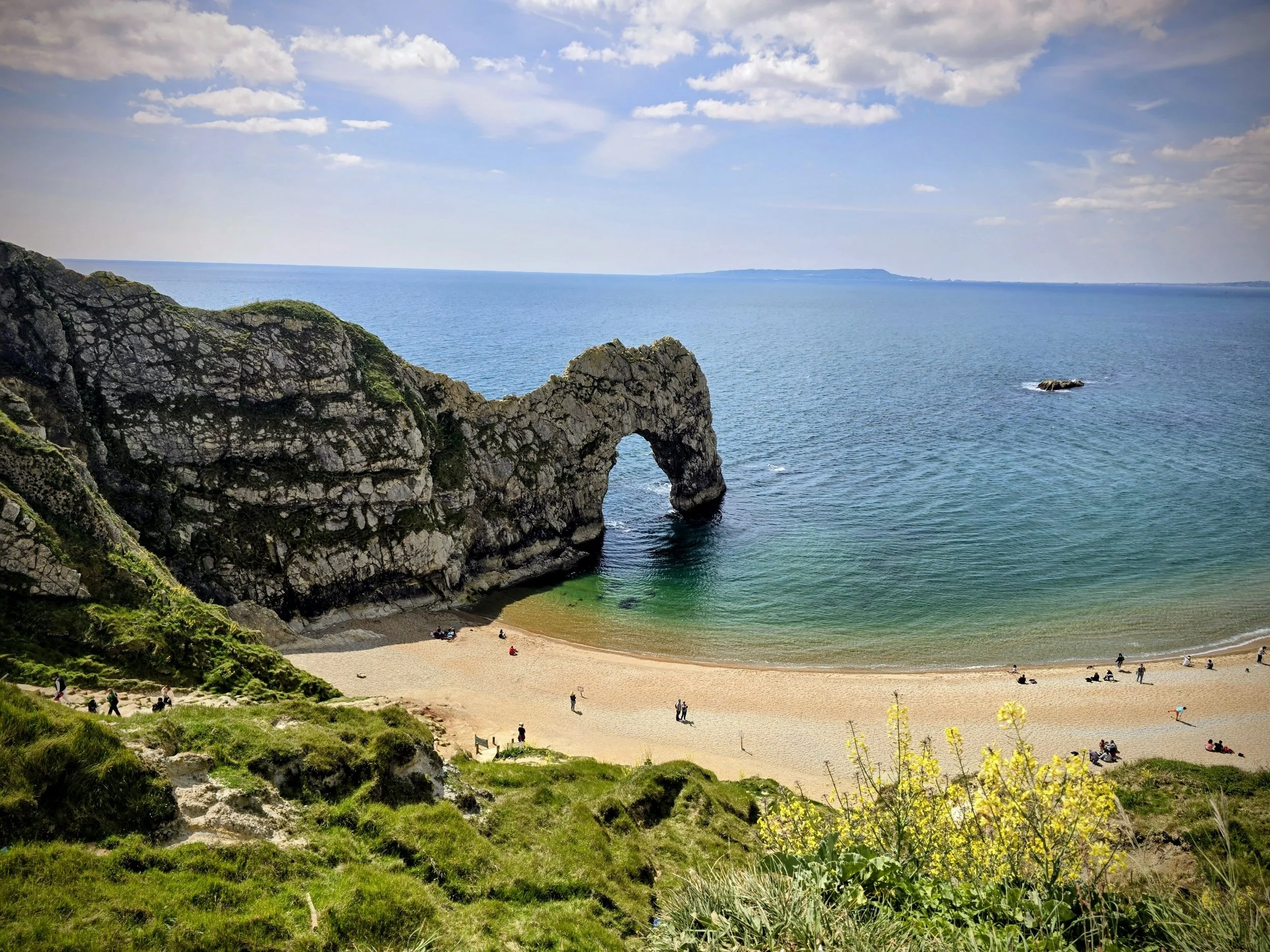 “Distant panoramic scene of Durdle Door with the iconic rock arch framed by rugged cliffs and a sweeping bay, waves gently meeting the pebble beach along the dramatic Jurassic Coast landscape.”