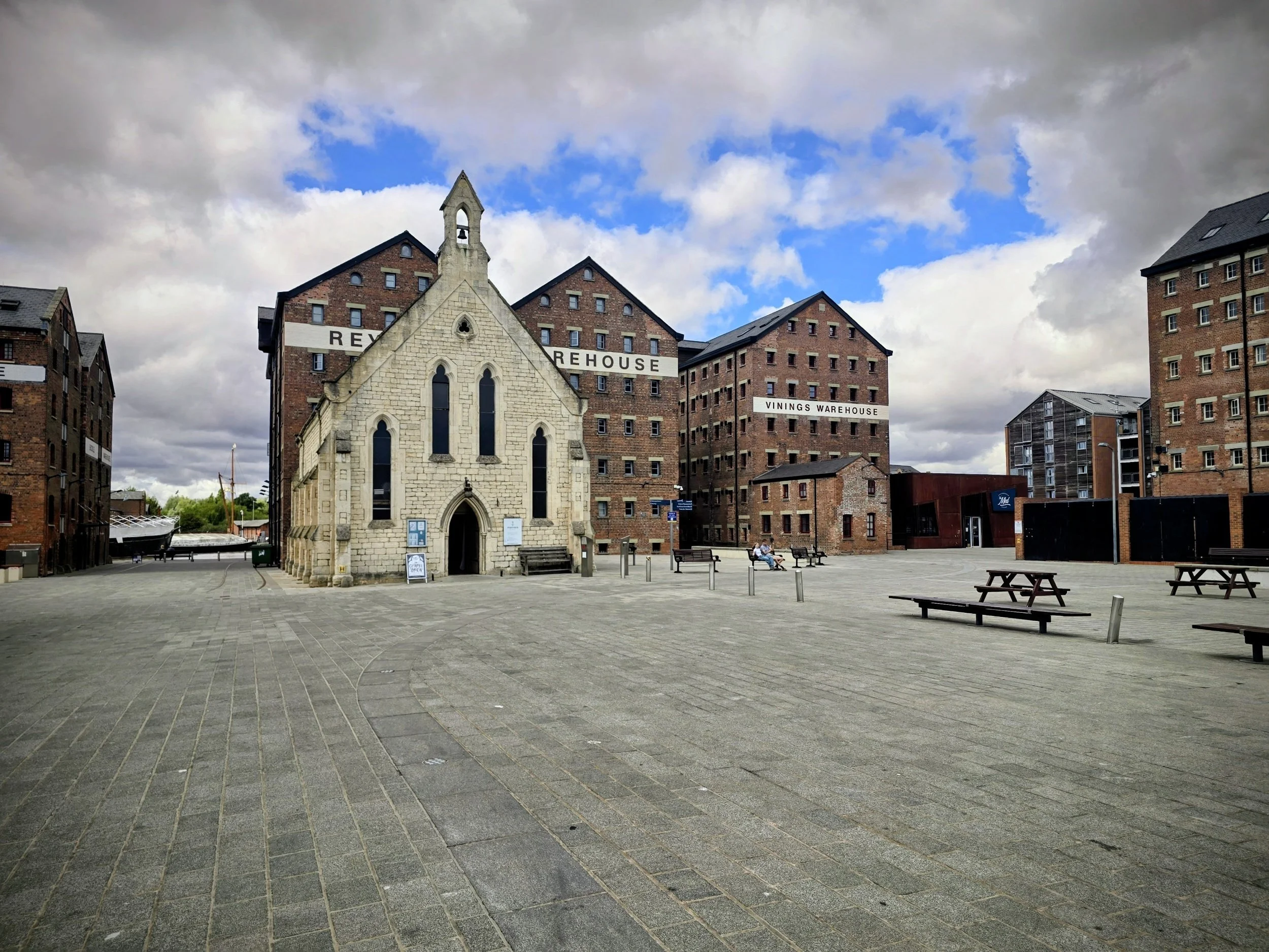 Open square with a small stone church in the foreground and modern brick warehouse buildings in the background, under cloudy sky with patches of blue.