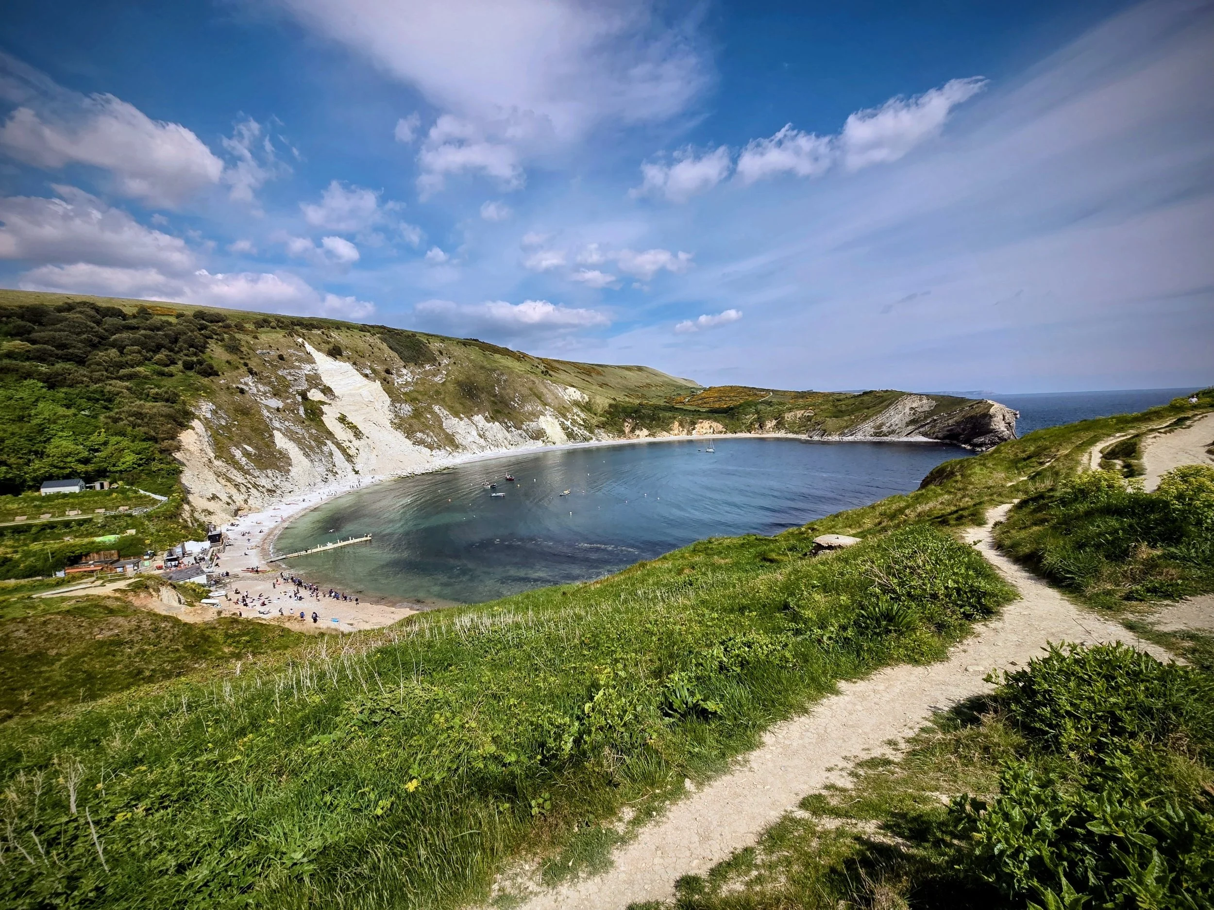 “Panoramic view of Lulworth Cove showing the perfectly curved bay with turquoise water, white chalk cliffs, and small fishing boats resting in the sheltered natural harbor.”