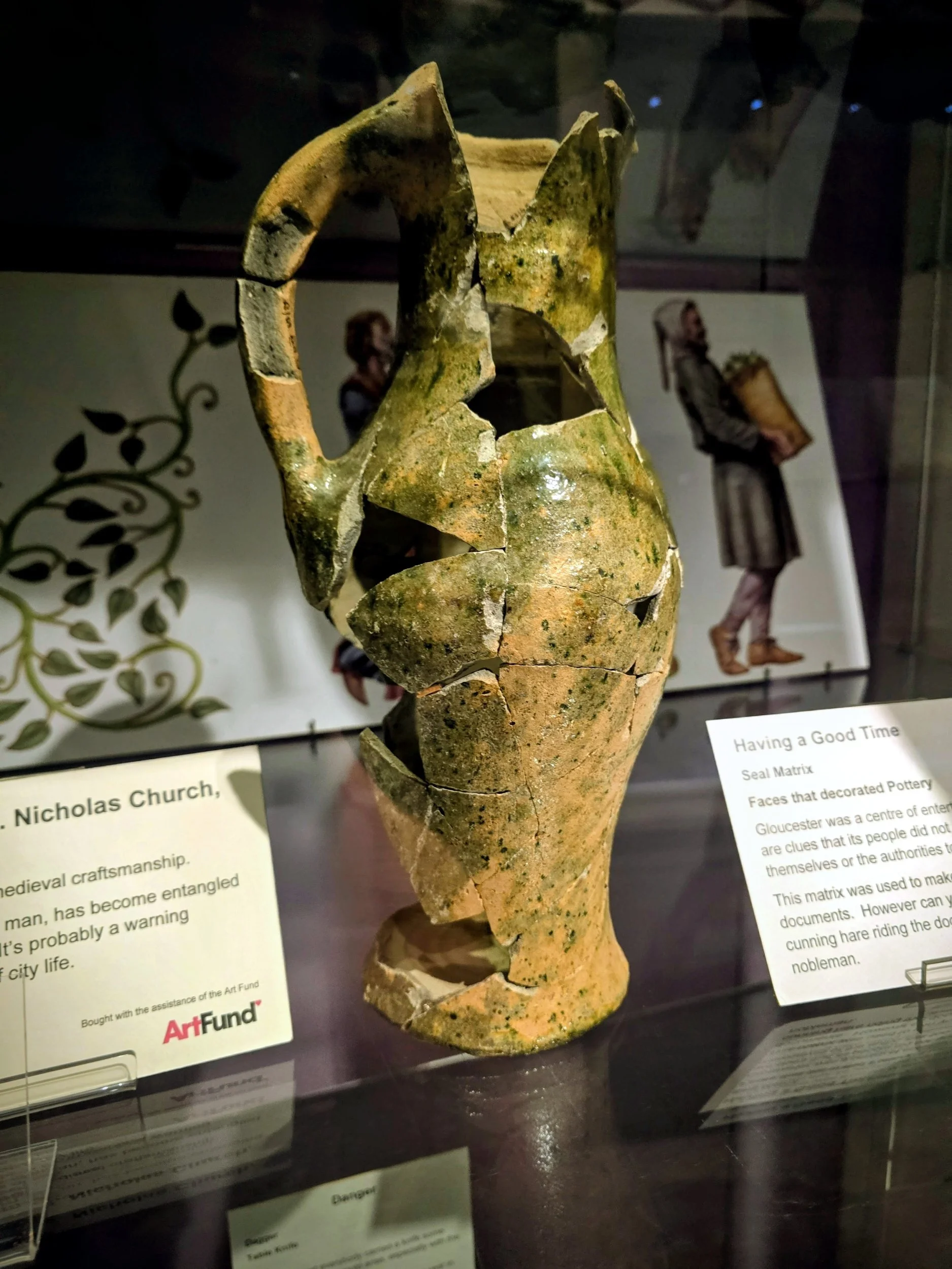Broken pottery mask with greenish and yellowish mossy surface displayed in a glass case at a museum exhibit.