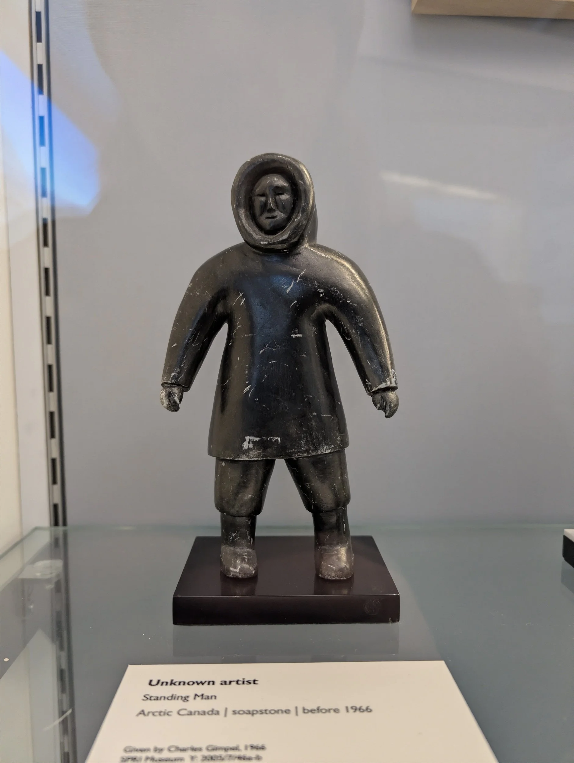 A soapstone sculpture of a standing person wearing a hooded coat and pants, with a neutral facial expression. The sculpture is displayed on a black base and is titled "Standing Man" by an unknown artist, created in Arctic Canada before 1966.