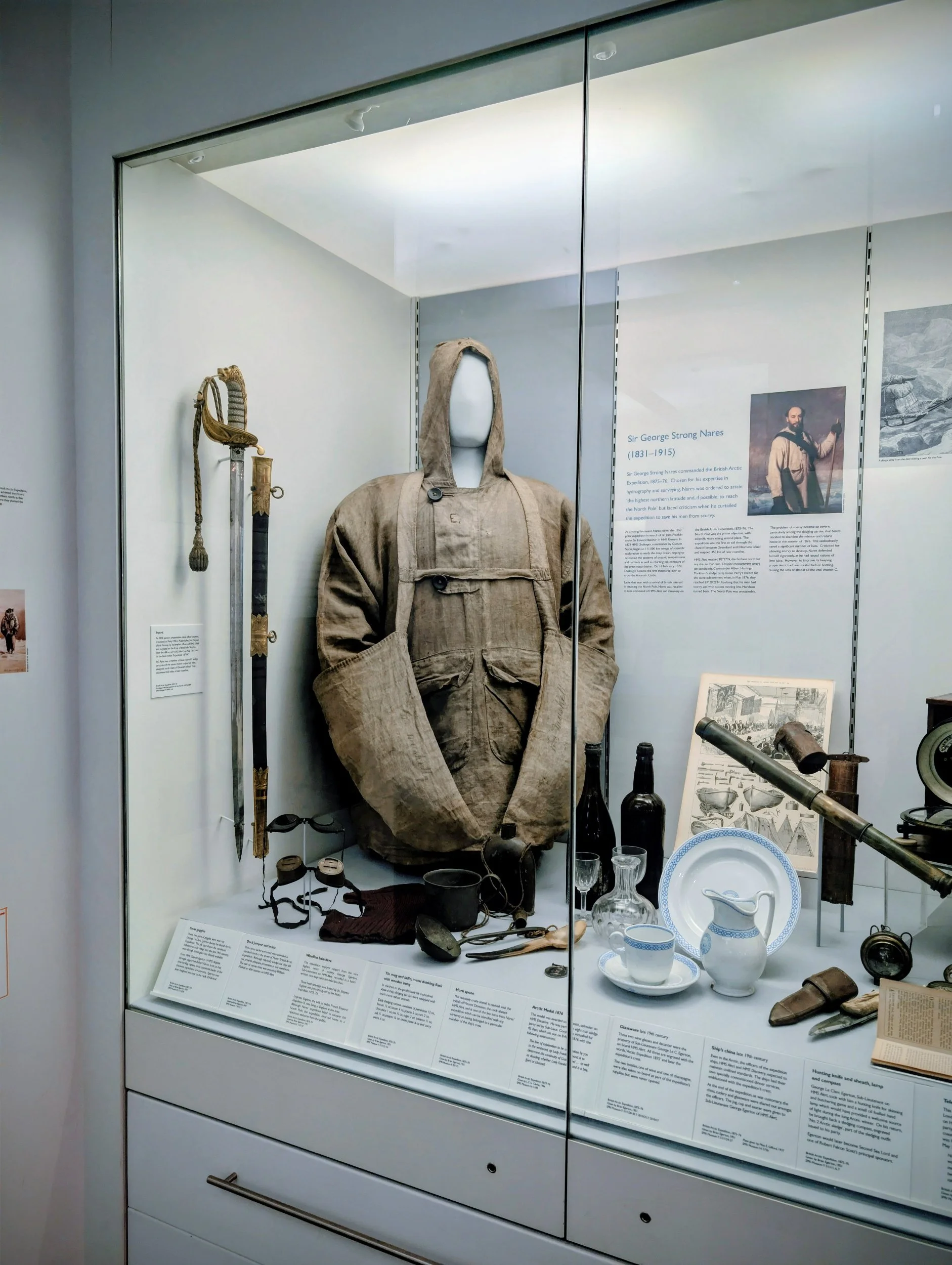 Historical museum display featuring vintage military gear including a beige coat with a hood, a sword, various bottles, a telescope, ceramics, and informational plaques. A portrait of Sir George Strong Nares is visible in the background.