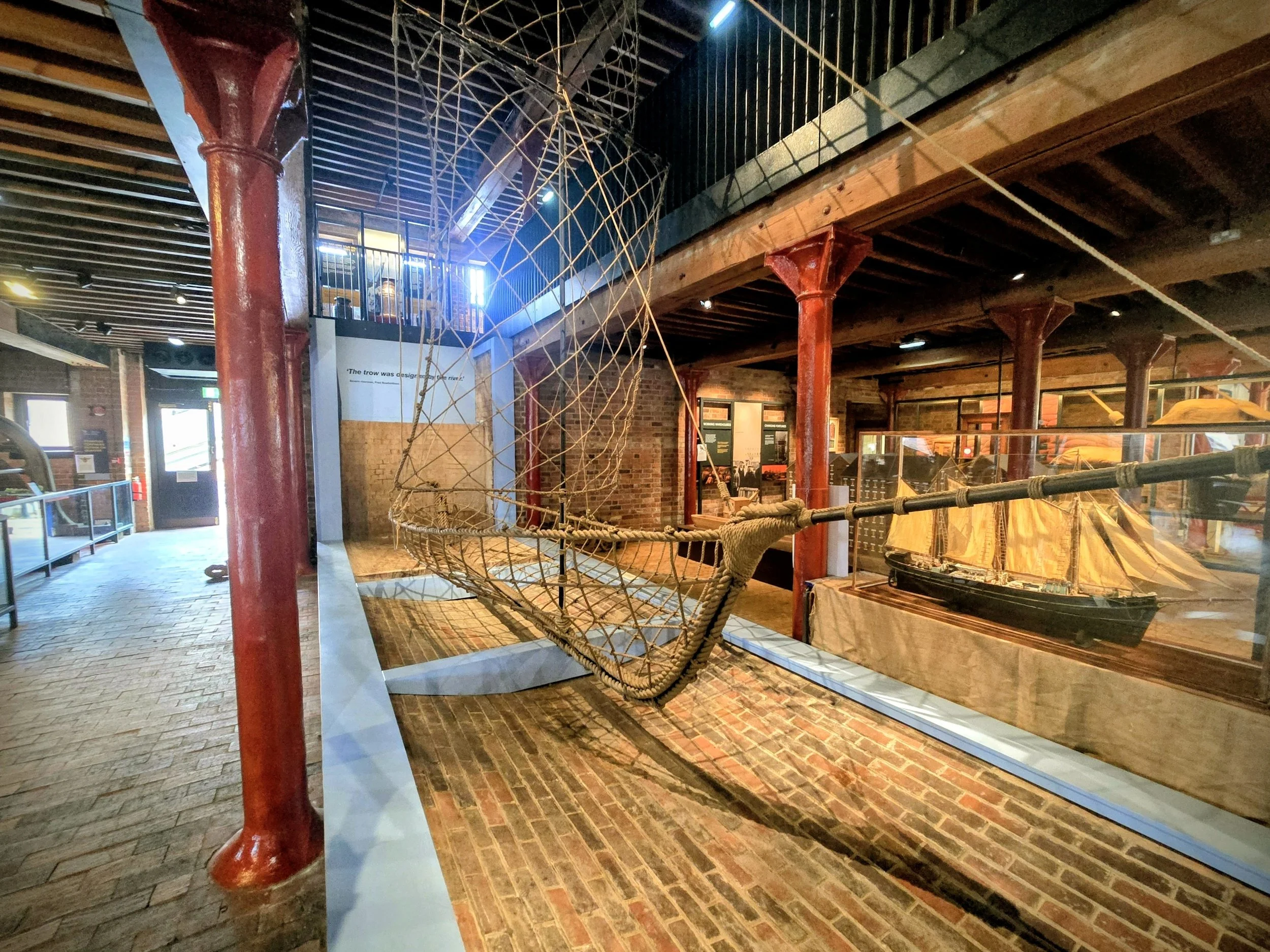 Historical maritime museum exhibit featuring a large wooden tugboat and a sailing ship model displayed behind glass, with a mesh fishing net hanging above.