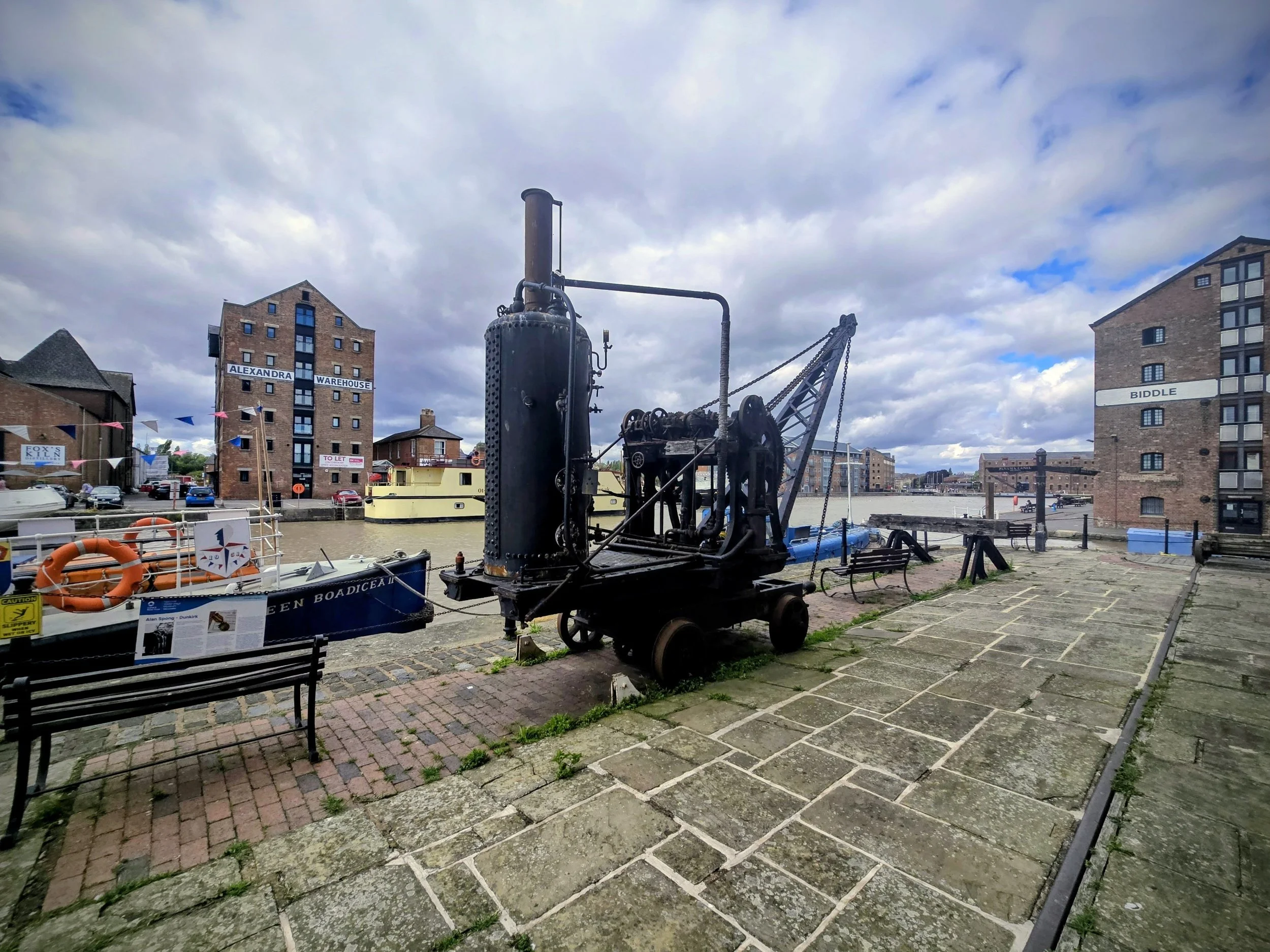 Historical steam-powered machine on a waterfront promenade with modern apartment buildings in the background, under a cloudy sky.