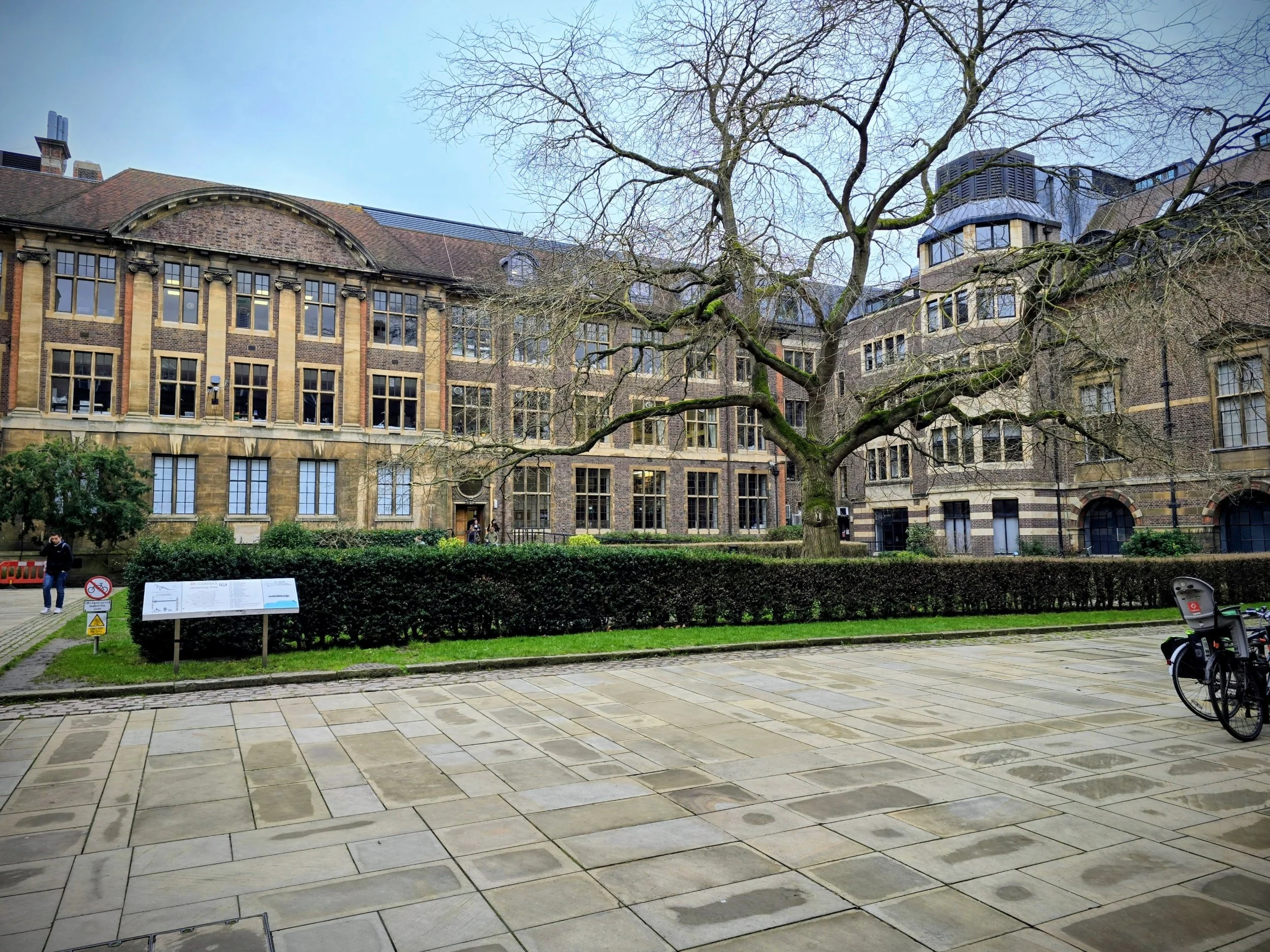 A historic brick building with large windows, a leafless tree in front, and a paved courtyard with a bicycle on the right. There are signs and a person walking on the left.