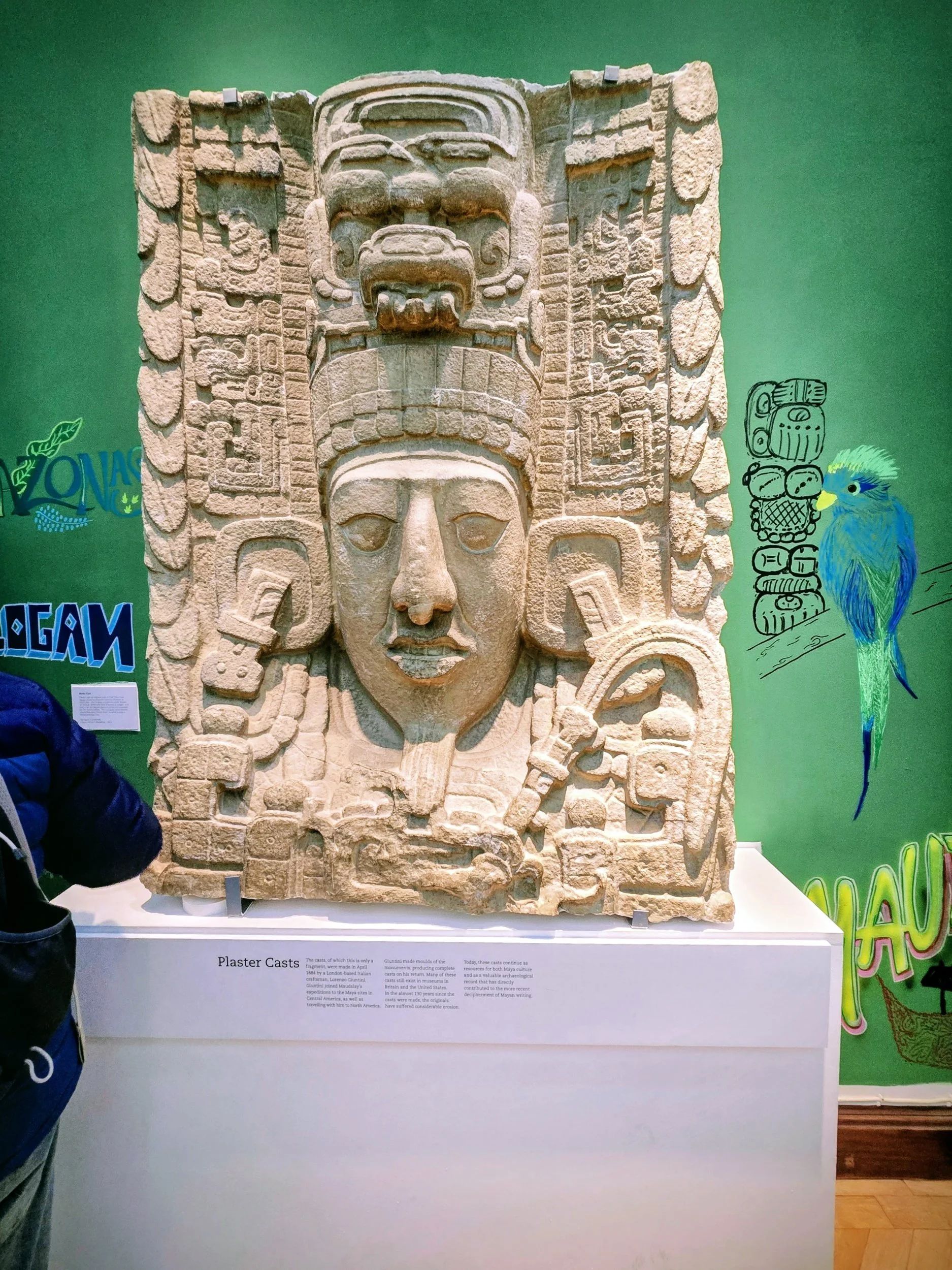 An ancient stone relief sculpture with a large human face, surrounded by intricate carvings and smaller figures, displayed in a museum setting.
