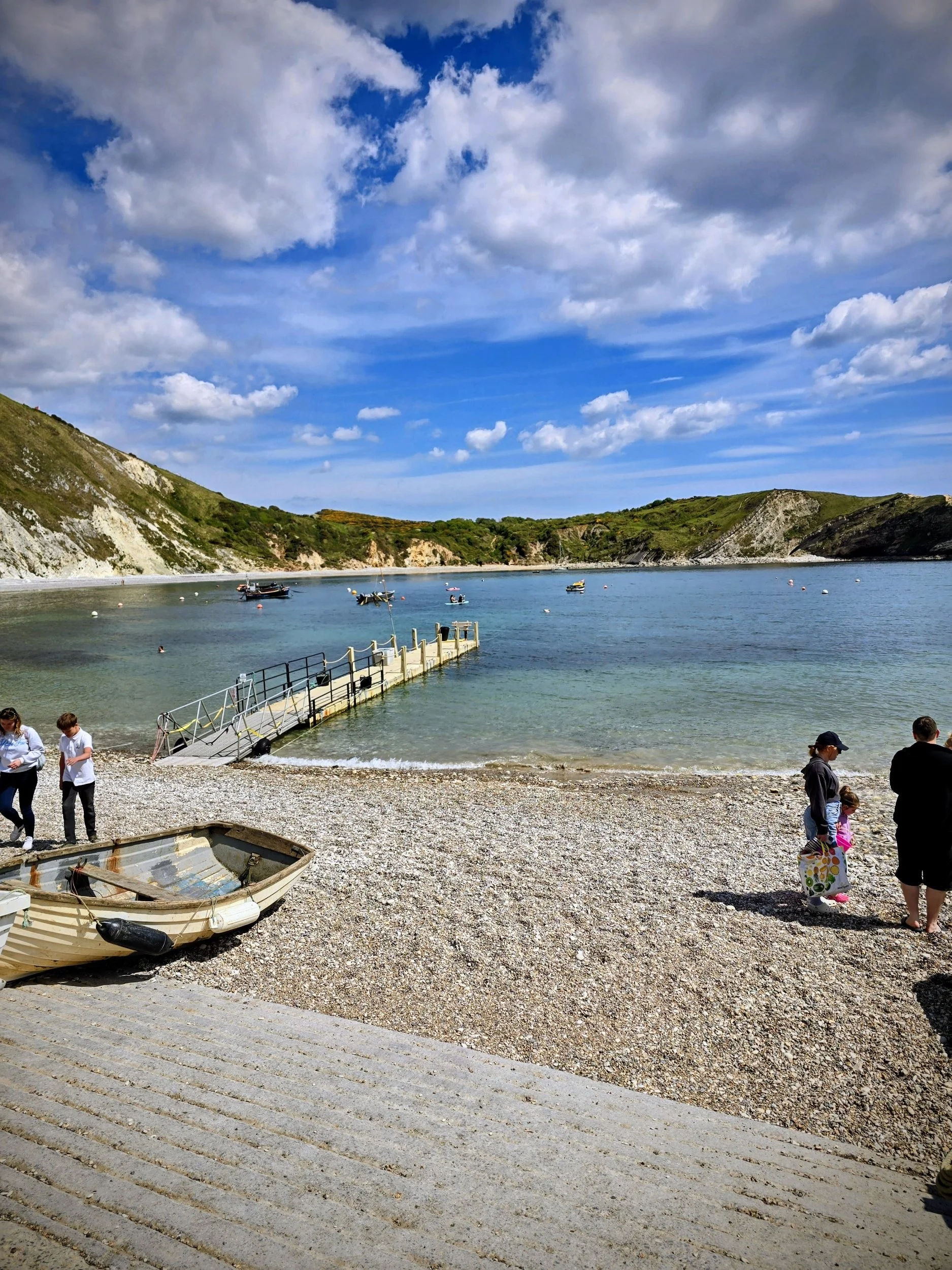 “View from the pebble beach at Lulworth Cove featuring calm clear water, curved limestone cliffs enclosing the bay, and small boats floating in the sheltered cove.”