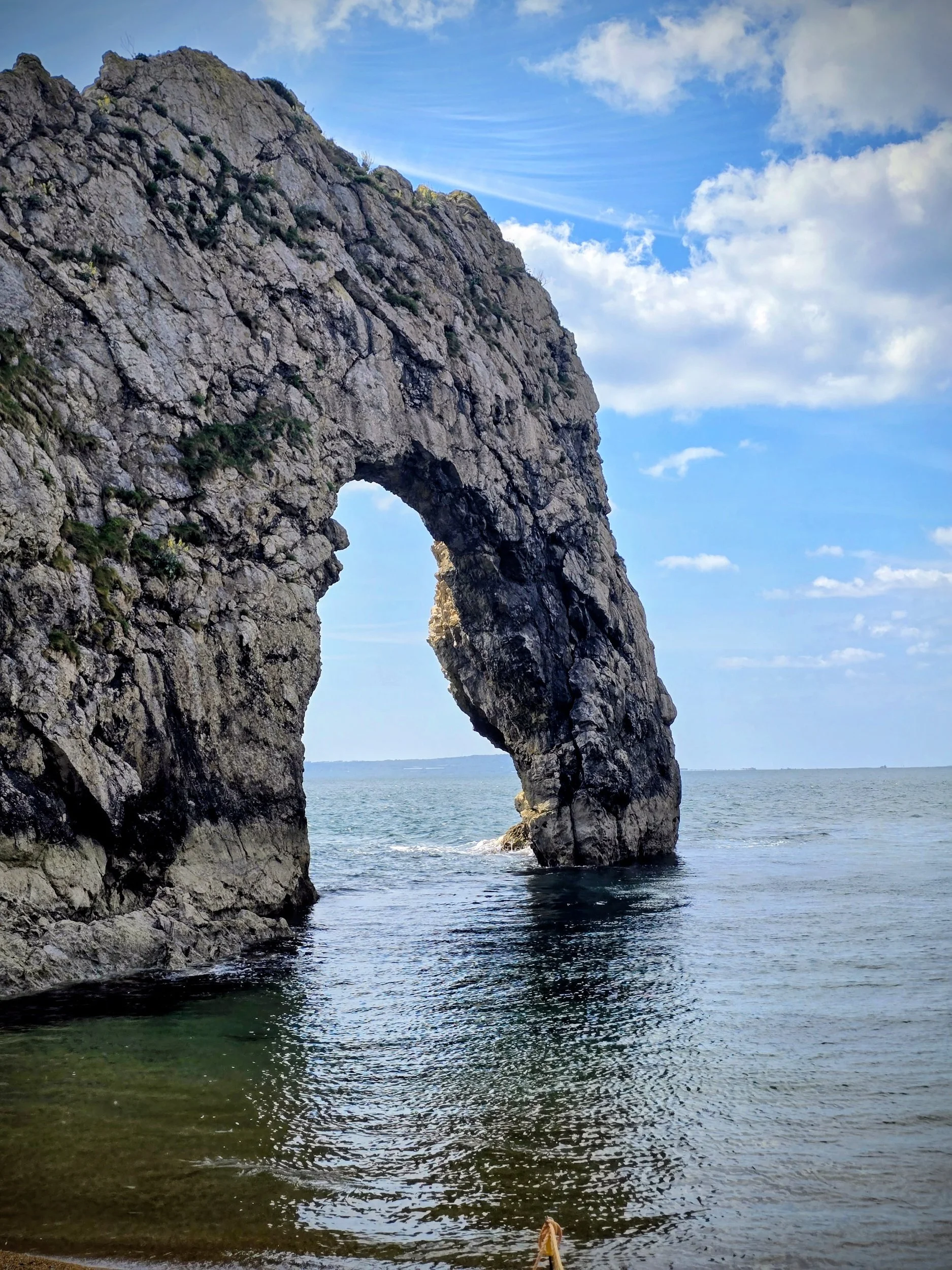 “Closer view of Durdle Door highlighting the massive limestone arch carved by the sea, clear blue water passing through the opening and textured rock surfaces visible against the coastal backdrop.”