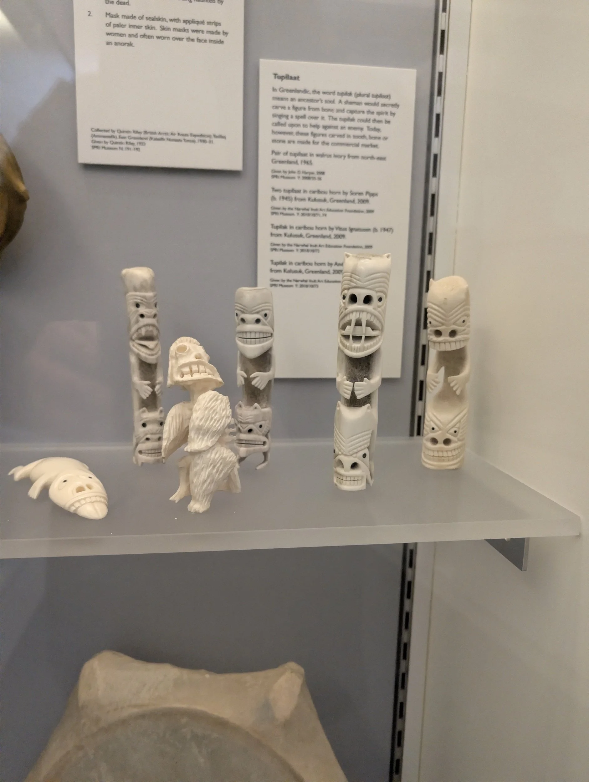 Collection of ivory-colored traditional Tiki artifacts with carved faces, including Tiki masks and figures, displayed on a gray shelf at a museum exhibit.