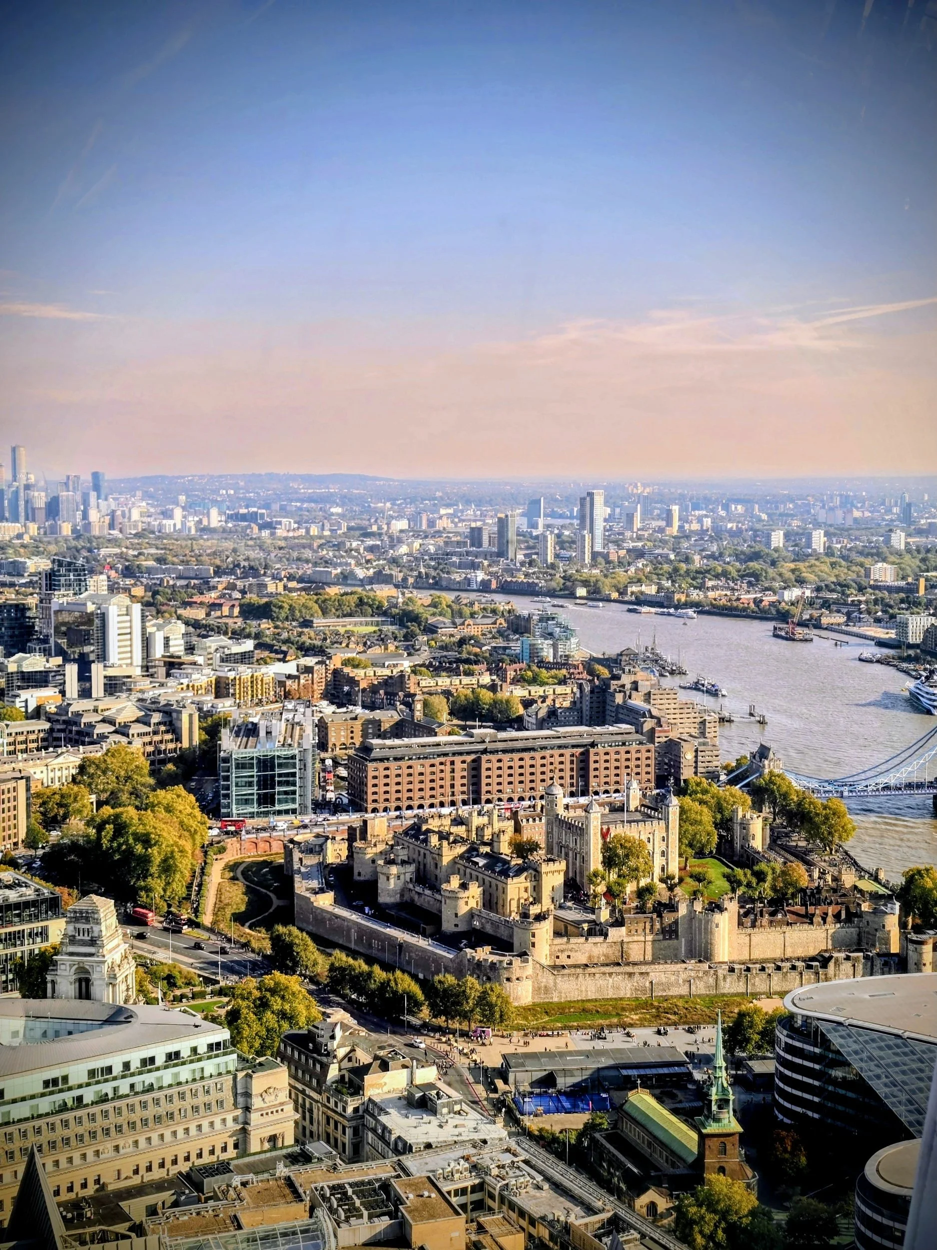 Aerial view of London with the Tower of London in the foreground and the River Thames winding through the city, surrounded by modern and historic buildings, under a clear sky.
