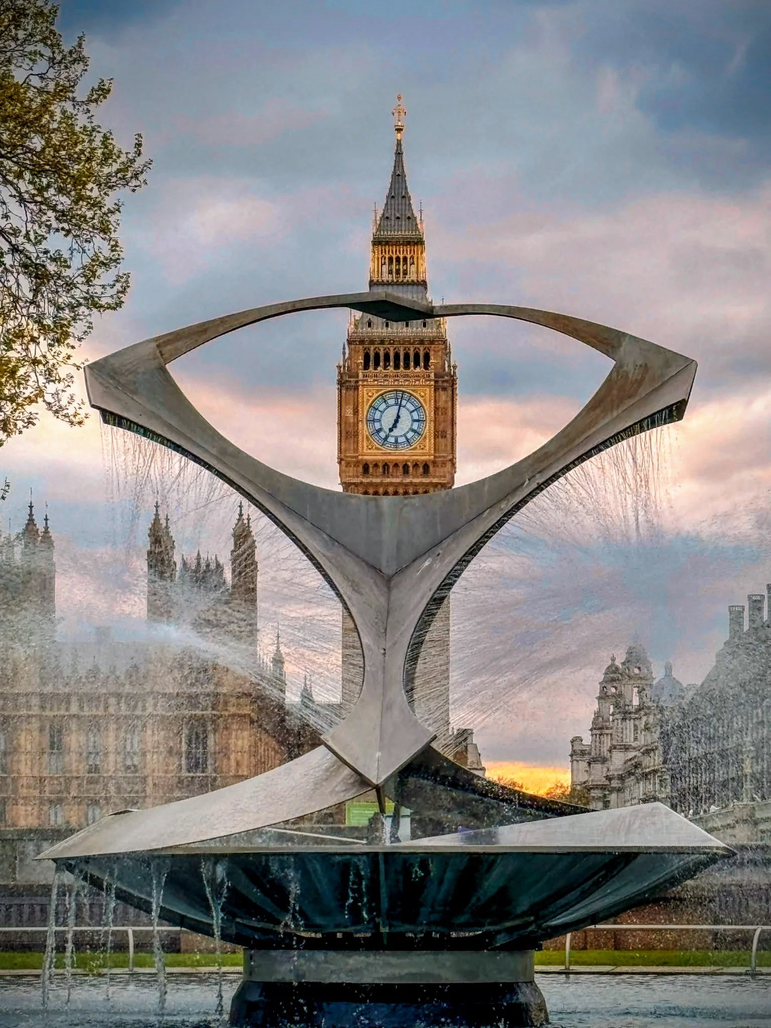 A modern metallic fountain sculpture with water streams, with Big Ben and the Palace of Westminster in the background, during sunset in London.