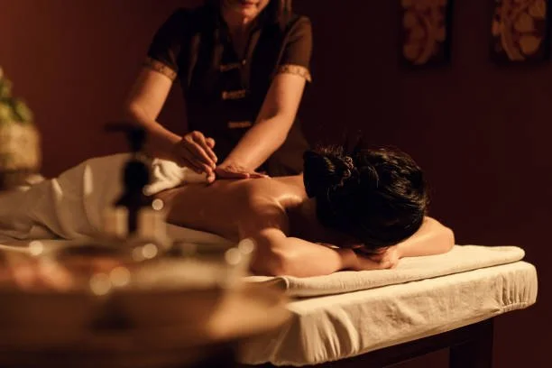 A woman is receiving a massage in a dimly lit spa, lying face down on a massage table with her head turned to the side.