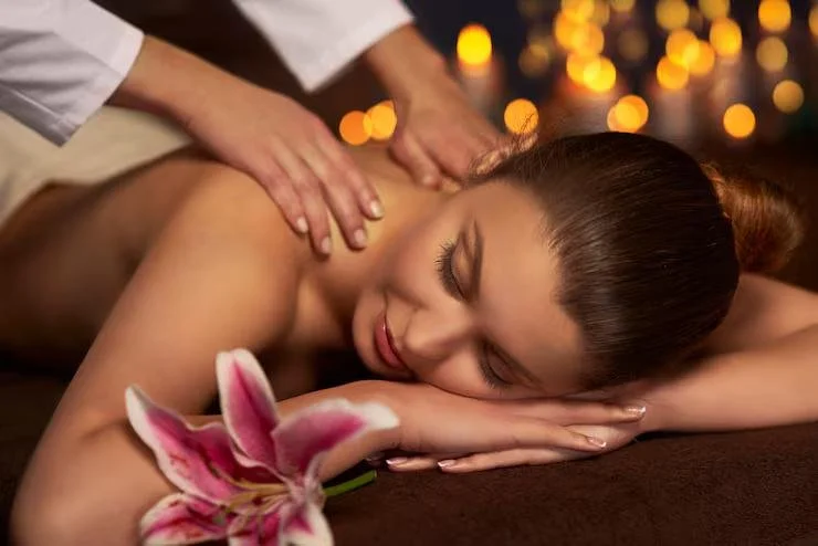 A woman receiving a massage with a pink lily flower nearby, in a serene setting with warm, blurred lights in the background.
