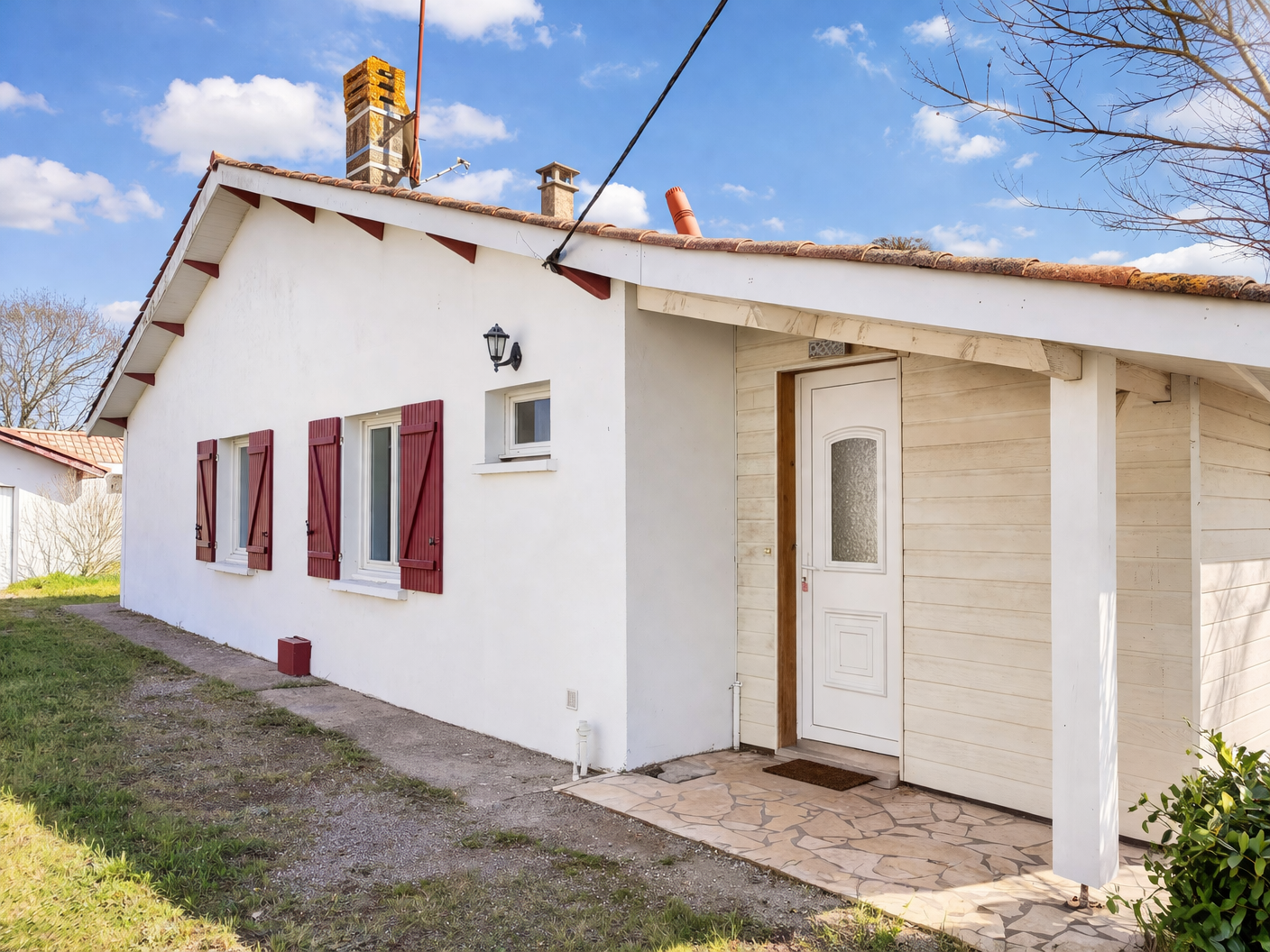 Vue de face d'une maison avec murs blancs, volets rouges aux fenêtres et une porte d'entrée blanche, sous un ciel bleu avec quelques nuages a cazaux