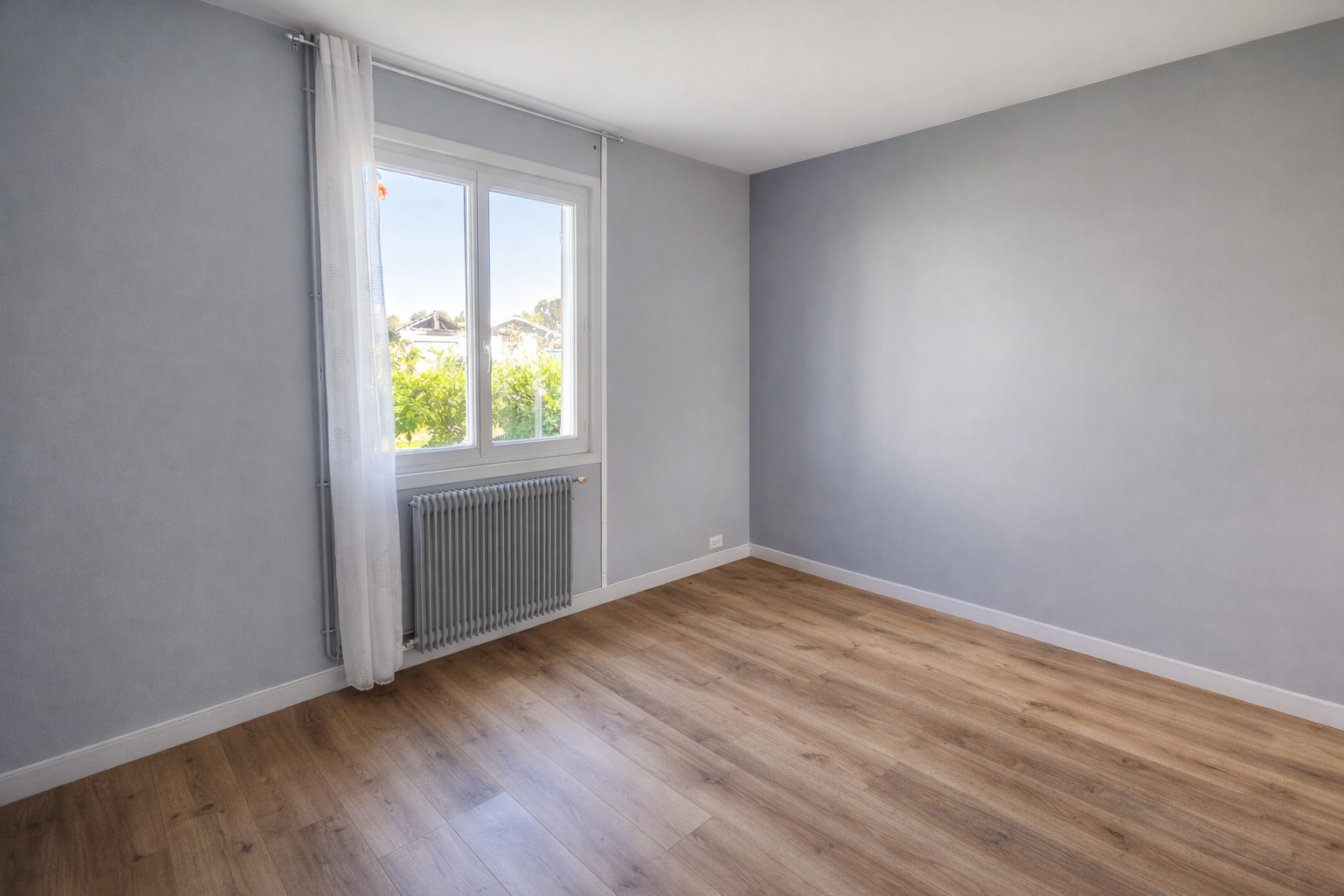 Chambre vide avec mur bleu clair, fenêtre avec rideau blanc, radiateur en dessous, sol en bois, vue sur un jardin avec des arbres et des maisons à cazaux