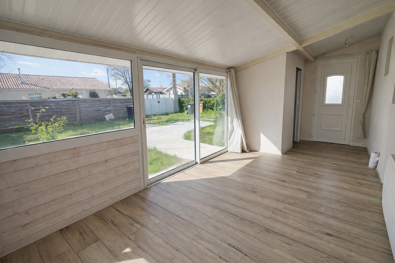 Intérieur d'une pièce avec de grandes fenêtres donnant sur un jardin, sol en bois clair, murs blancs, porte d'entrée blanche avec un petit vitrage, rideaux blancs à cazaux