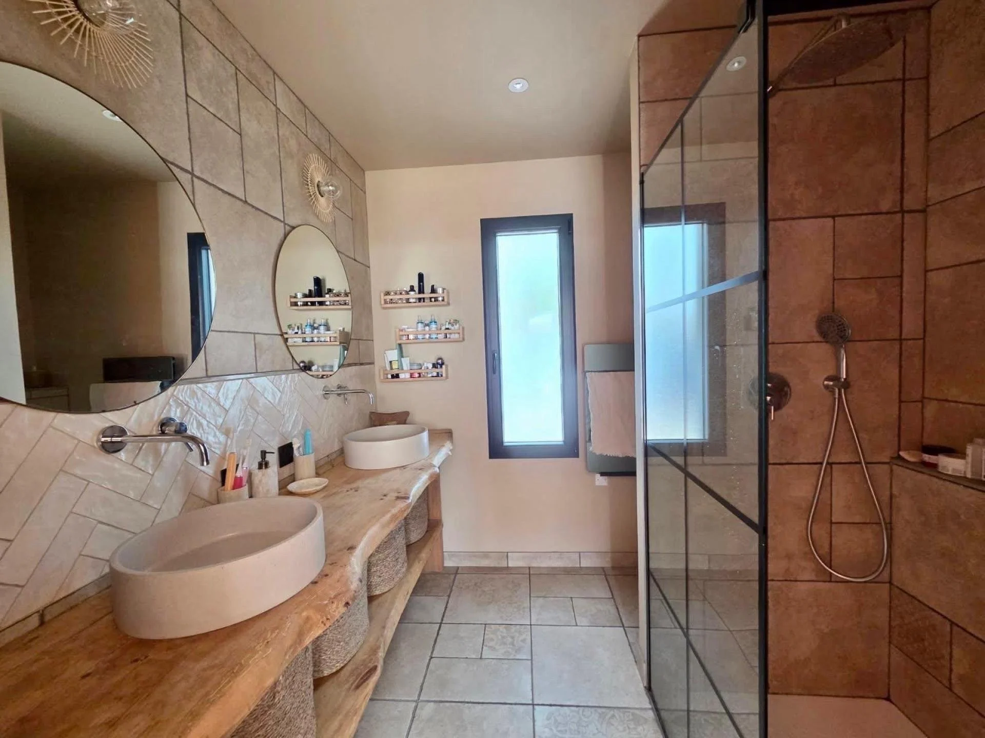 Salle de bain moderne avec deux lavabos ovales en pierre sur un comptoir en bois, grands miroirs ronds, une douche à l'italienne avec porte en verre, fenêtre rectangulaire, et étagères avec produits de toilette.