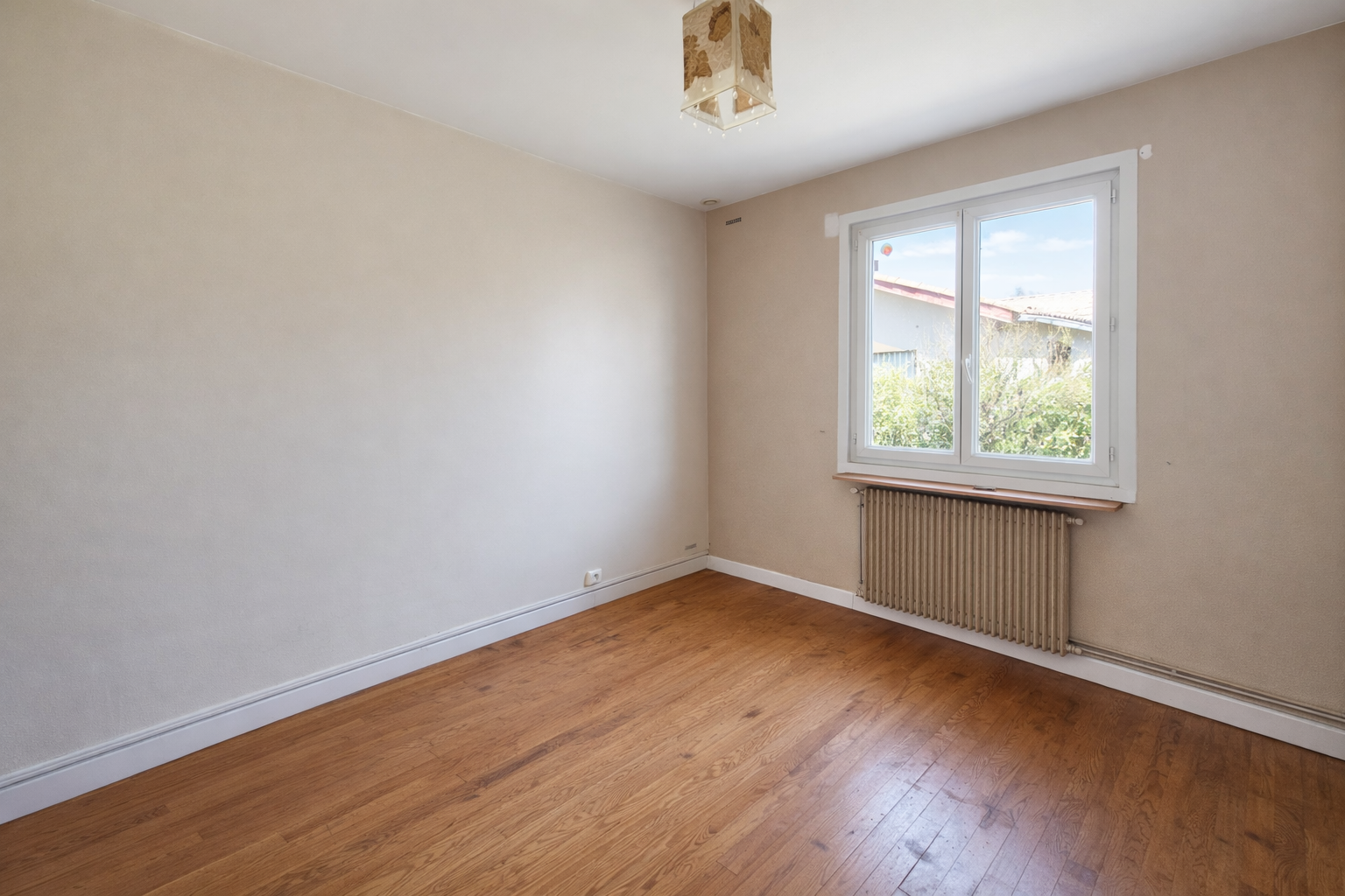 Pièce vide avec parquet en bois clair, mur beige, fenêtre blanche laissant entrer la lumière naturelle, radiateur en dessous de la fenêtre, plafond blanc avec un luminaire à cazaux