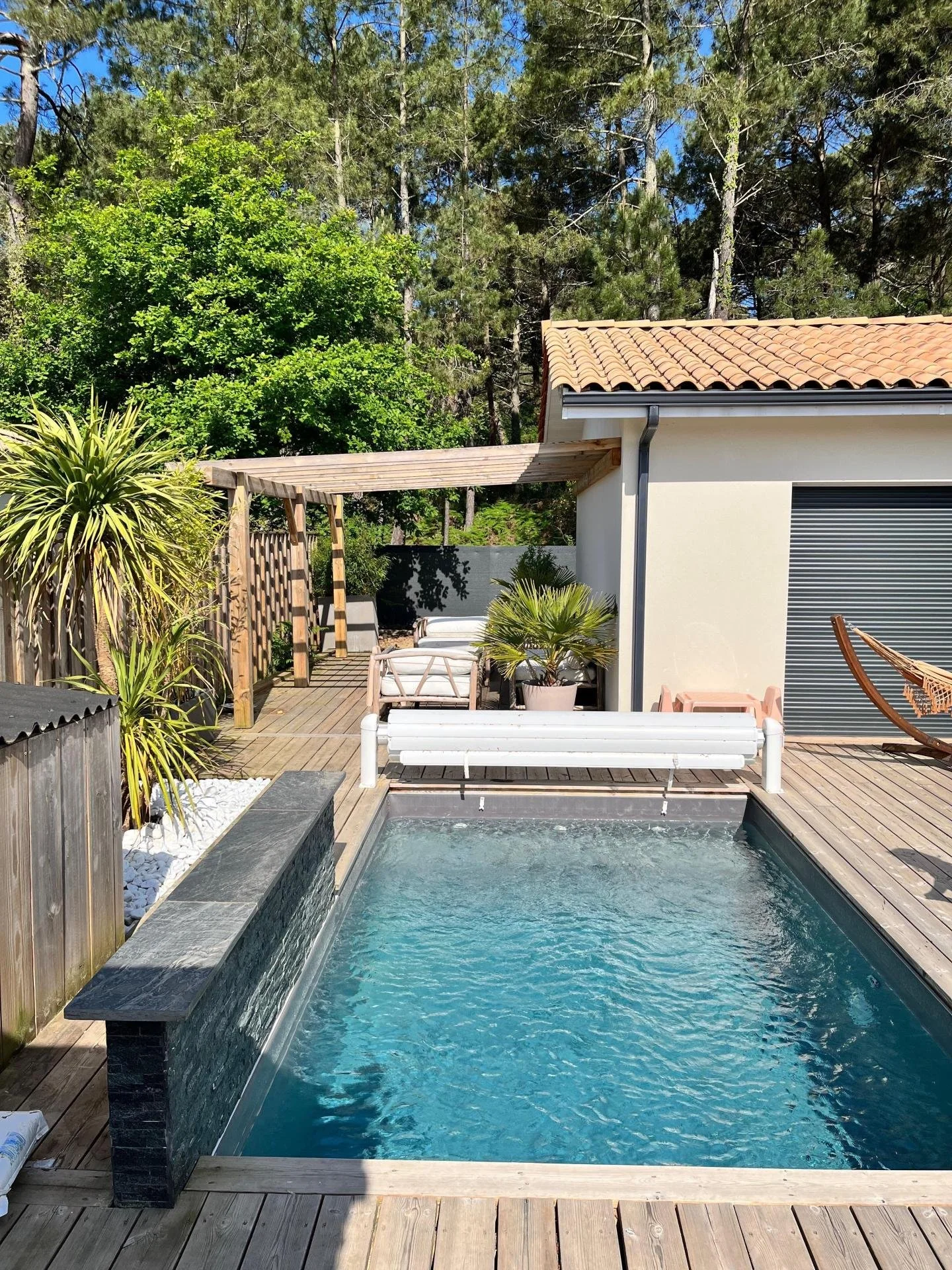 Piscine extérieure dans un jardin ensoleillé avec des plantes tropicales, une clôture en bois, un espace de détente avec un fauteuil et une pergola en bois, bâtiment avec toit en tuiles orange.