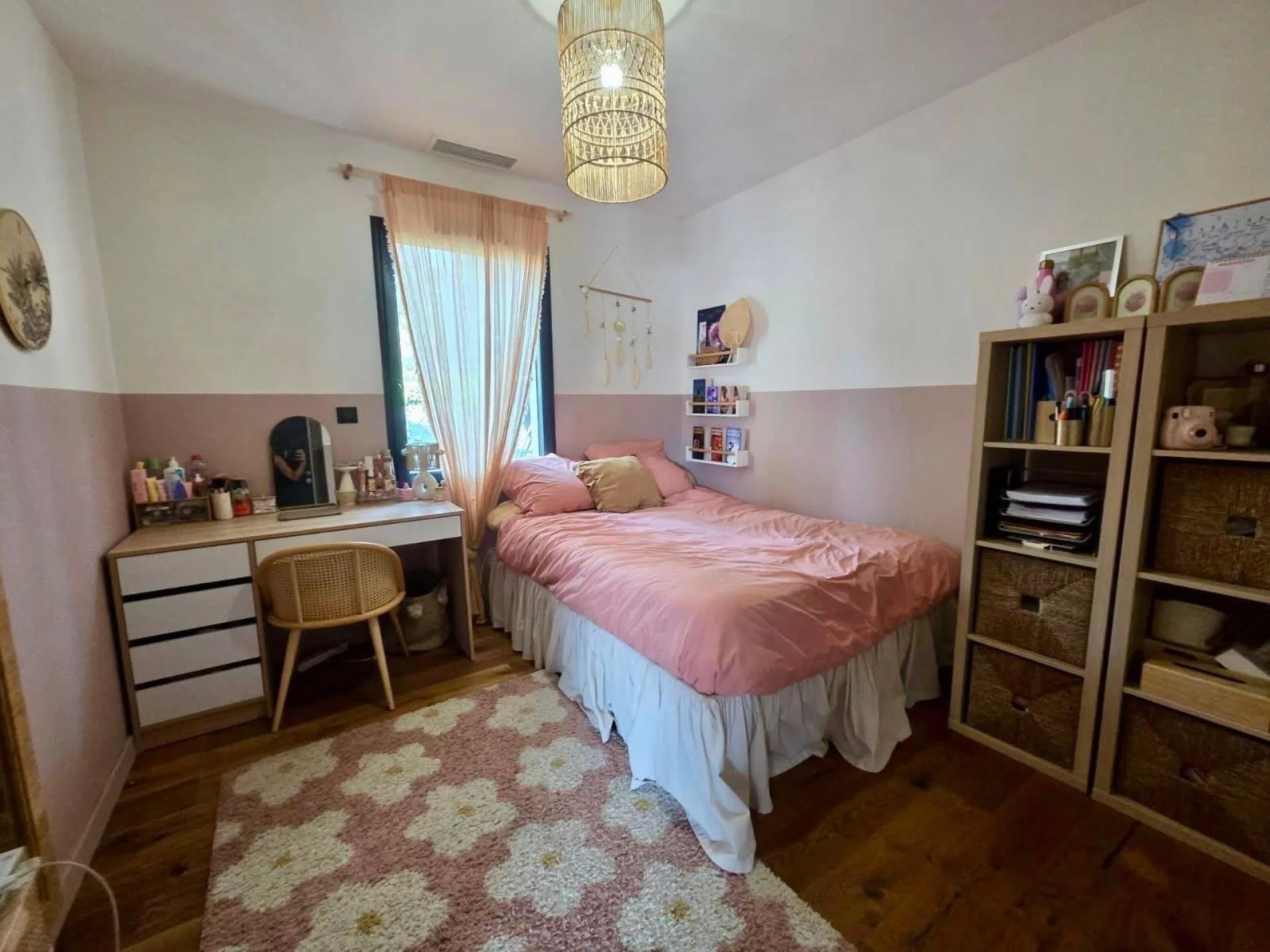 Chambre à coucher avec un lit rose, une fenêtre avec des rideaux, une étagère, un bureau avec un miroir, une armoire avec des paniers de rangement, un tapis floral, décoration bohème, mur deux tons, sol en bois.