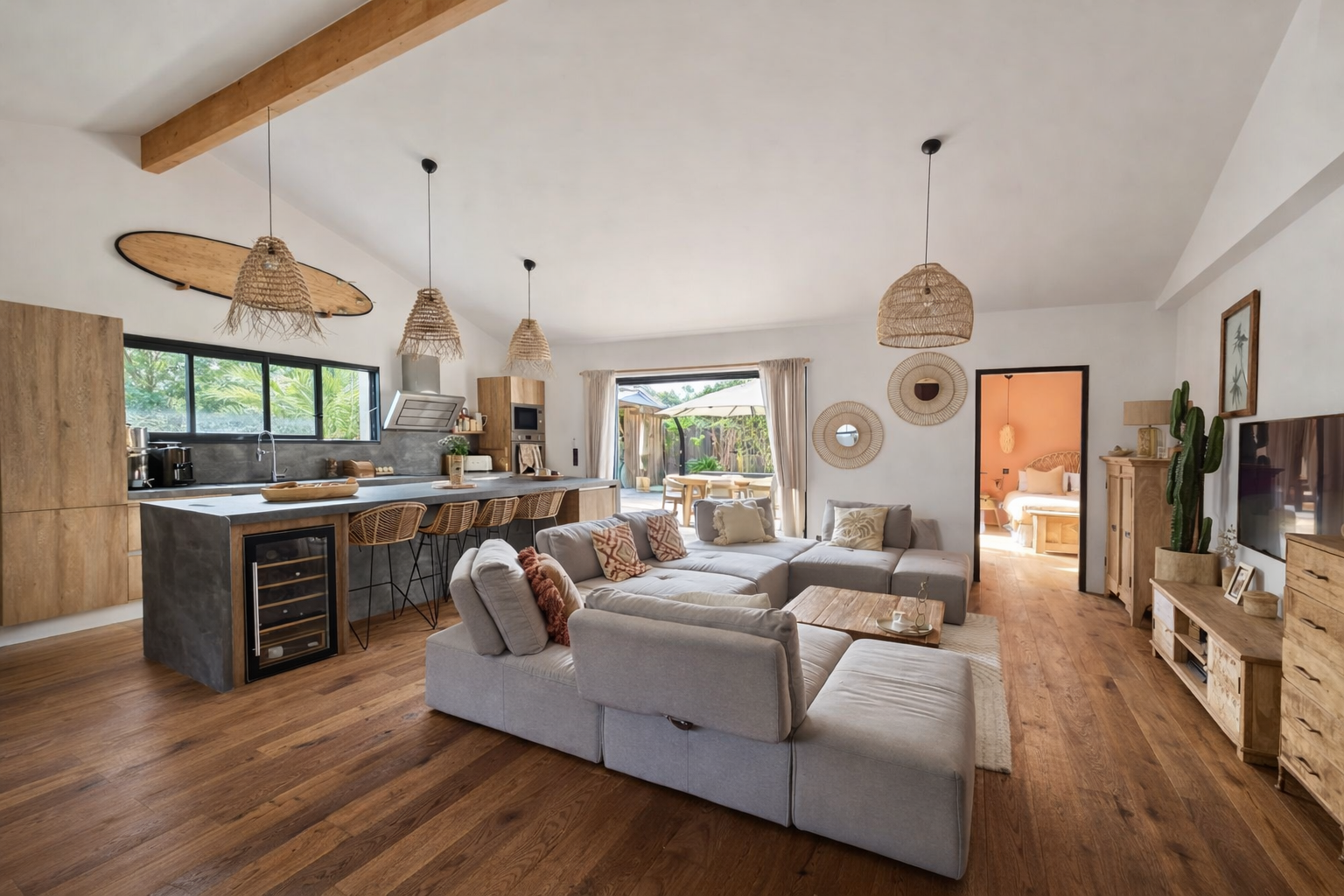 Salon et cuisine ouverts avec mobilier en bois, canapé en gris clair, décor naturel, tapis beige, lampes en rotin suspendues, porte-fenêtre menant à un espace extérieur, chambre visible à travers une porte, style bohème