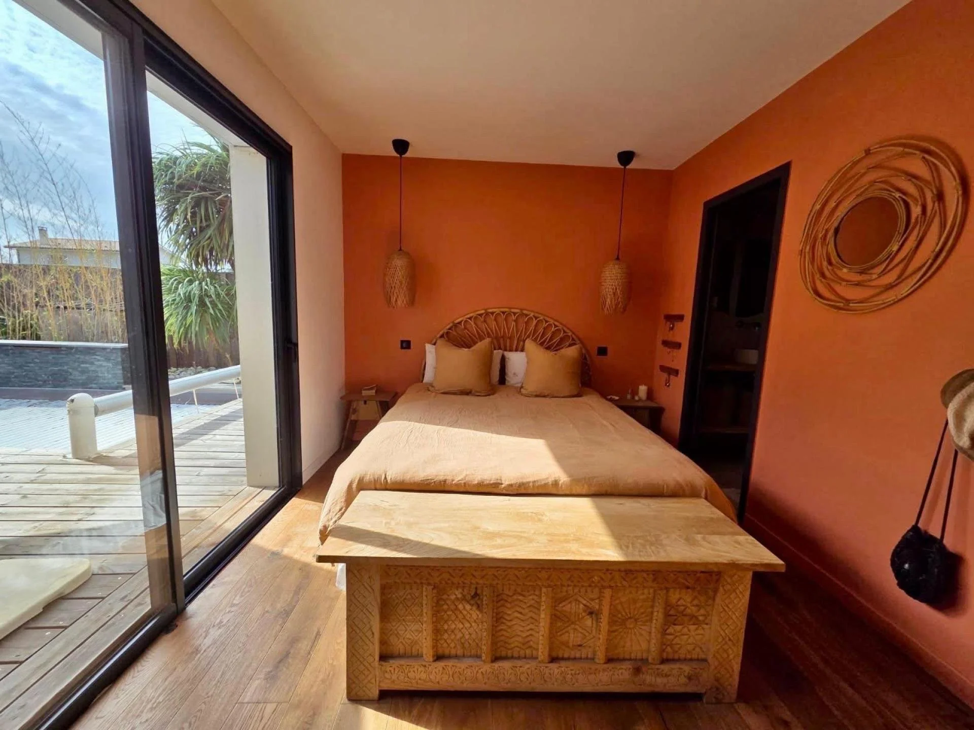 Chambre avec lit double, murs orange, décoration en bambou, grande fenêtre donnant sur un patio en bois avec plantes tropicales.
