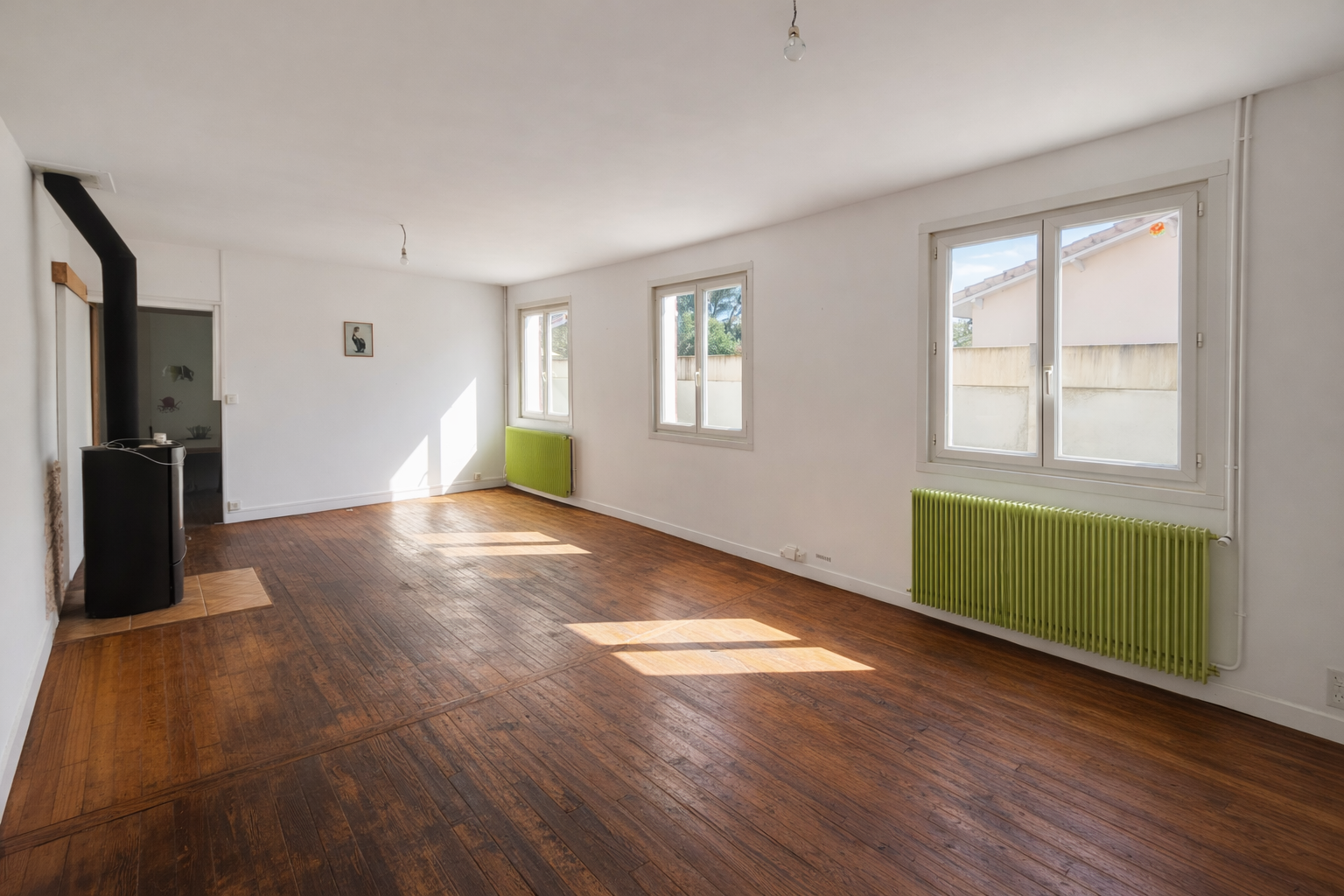 Grand salon lumineux avec trois fenêtres, parquet en bois, radiateurs verts et mur blanc à cazaux