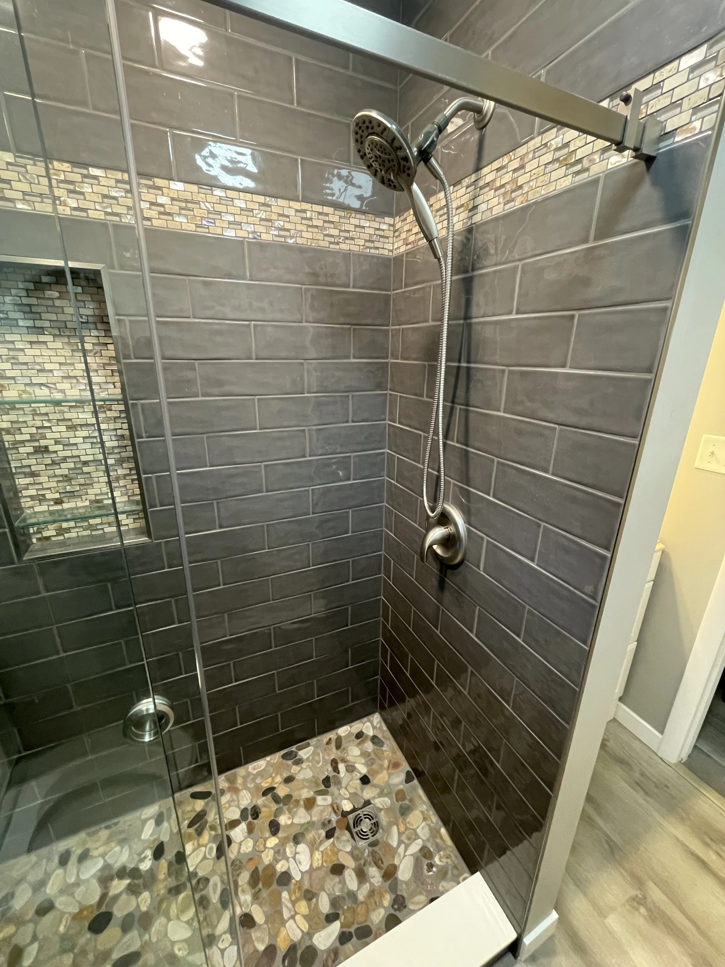 A modern shower with gray subway tile walls, a decorative mosaic tile border, a pebble tile floor, and a brushed nickel showerhead and fixture.