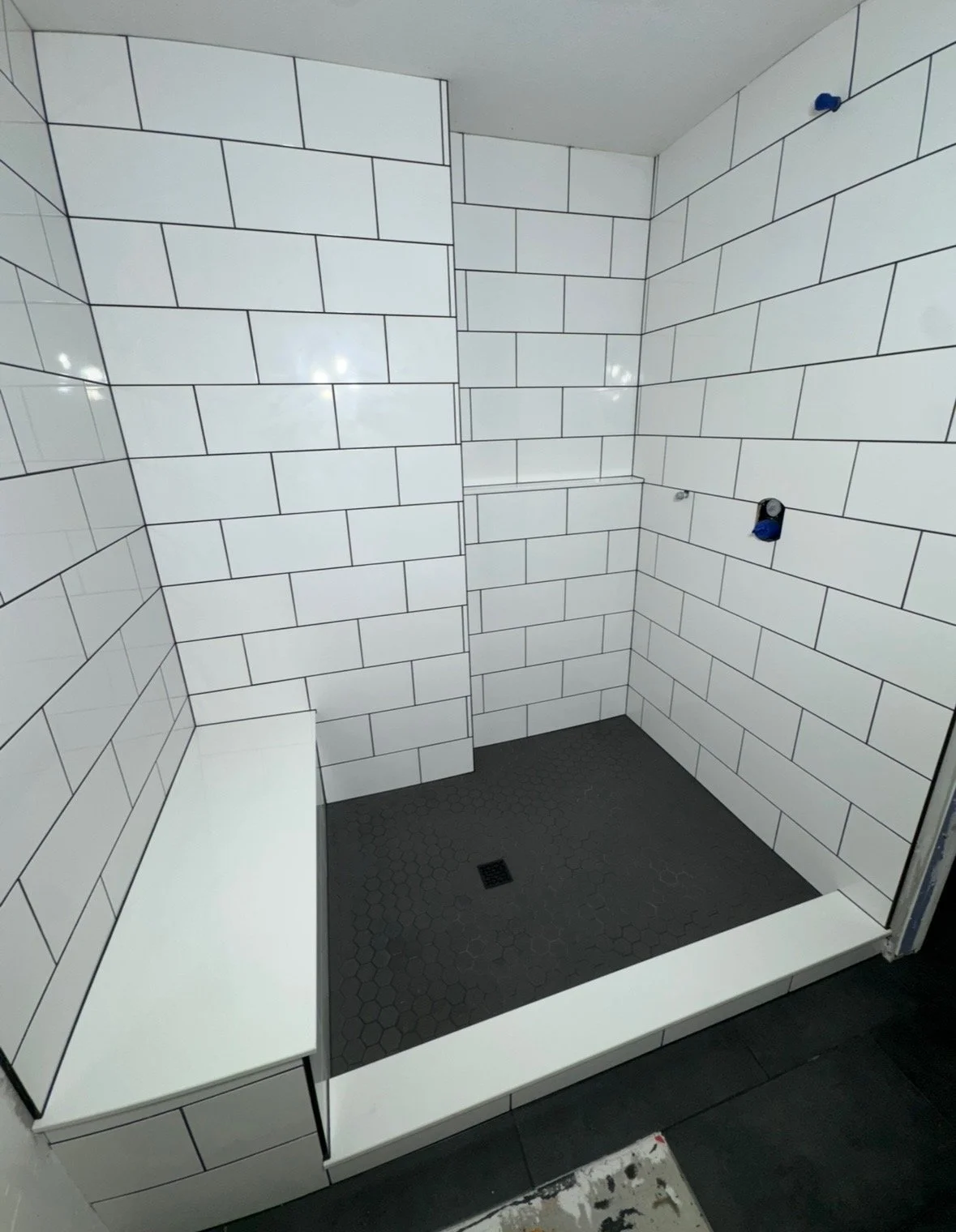Empty walk-in shower with white subway tile walls, black floor tiles with a drain, and a built-in bench on one side.