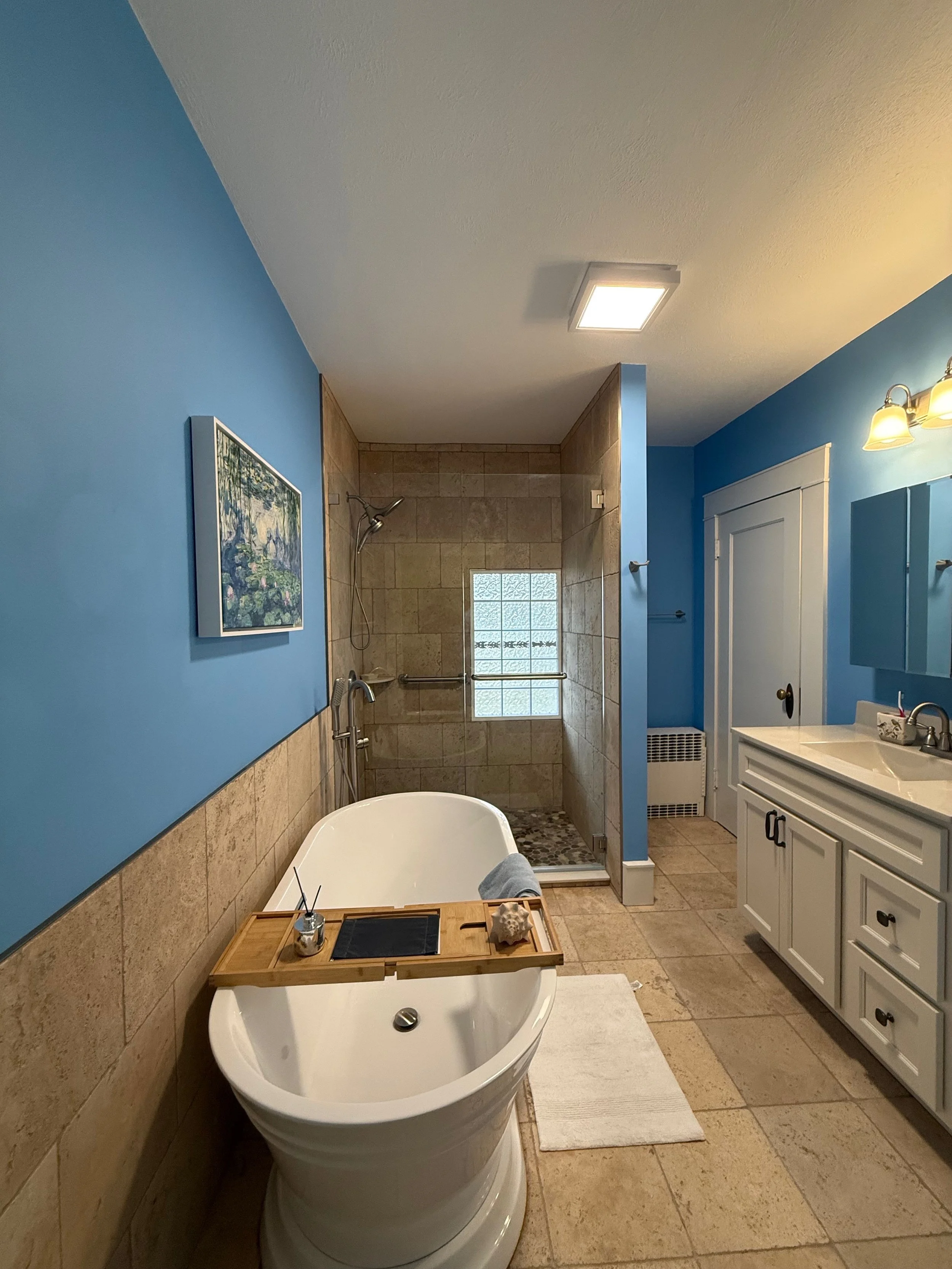 Bathroom with blue walls, a white freestanding bathtub with a wooden tray, a walk-in shower with beige tiles, a white vanity with a mirror, and a small window in the shower area.