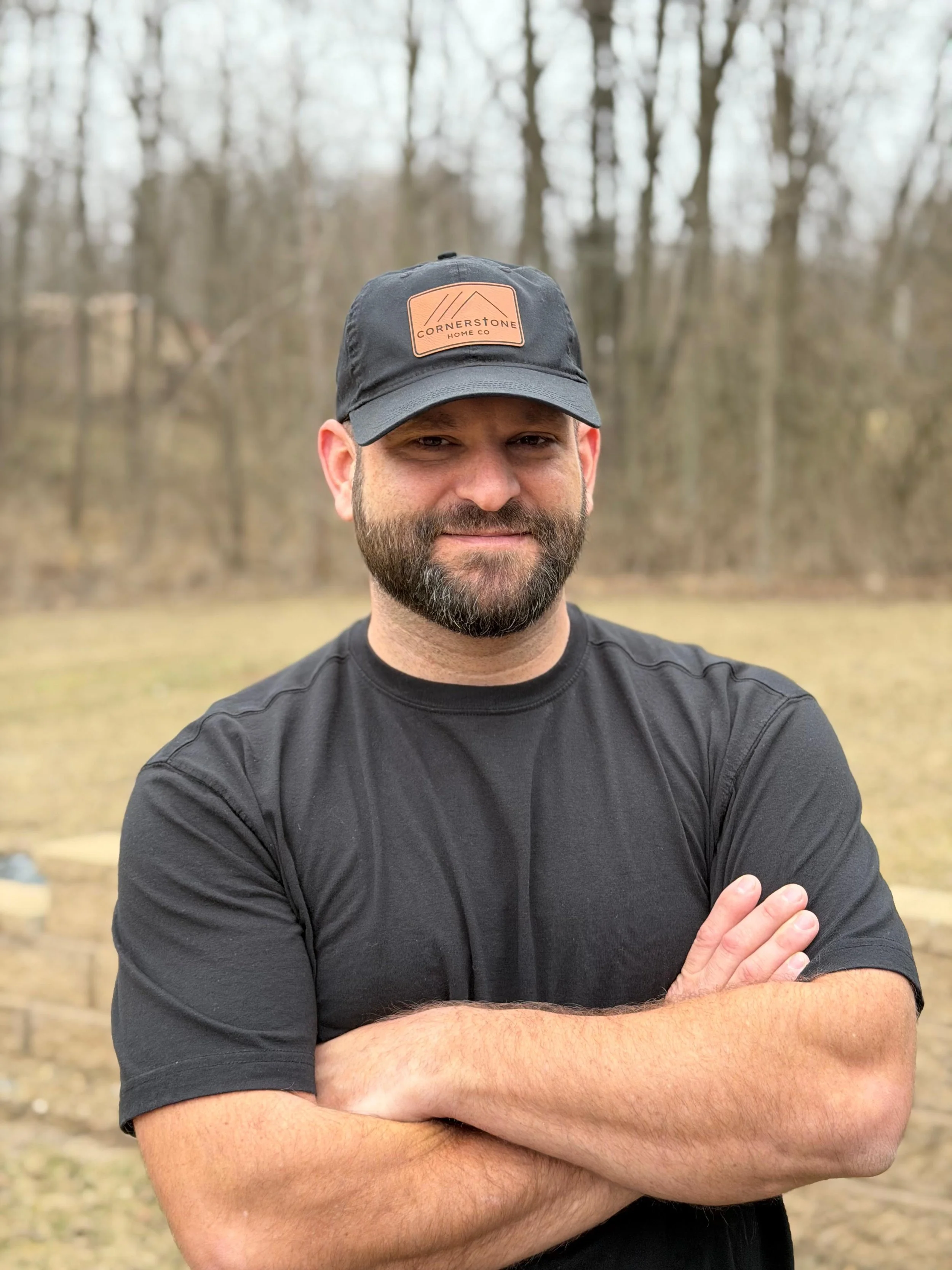 A man with a beard and short hair wearing a black cap with a tan logo and a black t-shirt, standing outdoors with a wooded background, arms crossed.