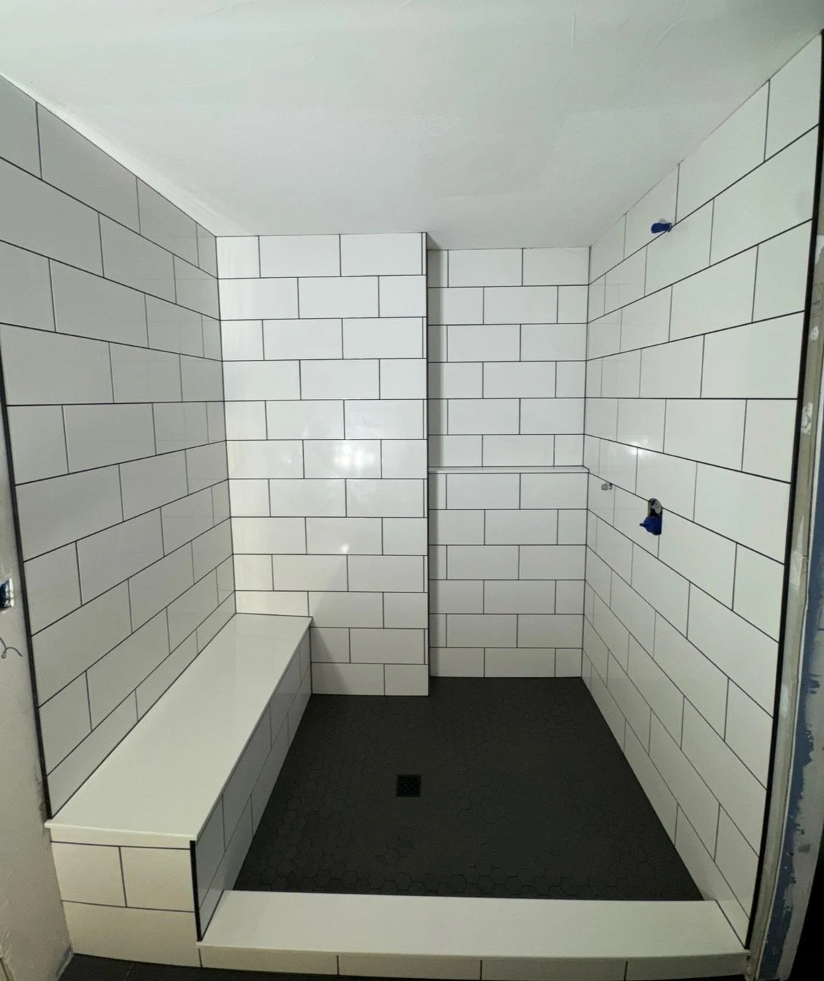Empty walk-in shower with white subway tiles, black hexagonal floor tiles, built-in bench, and blue fixtures.