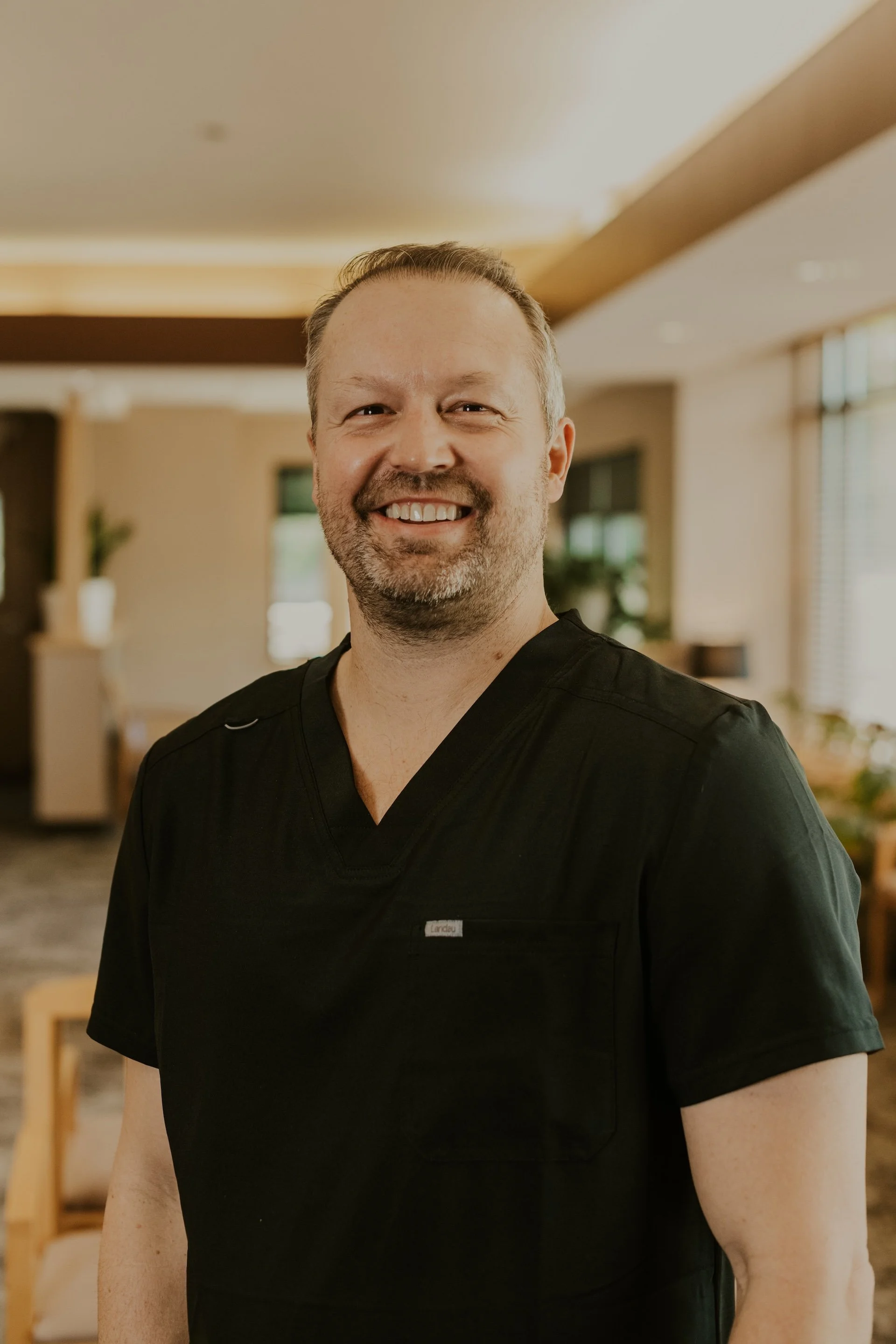 Dr. Chad Nelson smiling while wearing black scrubs and standing in the warmly lit Lakeville Family Dental office.