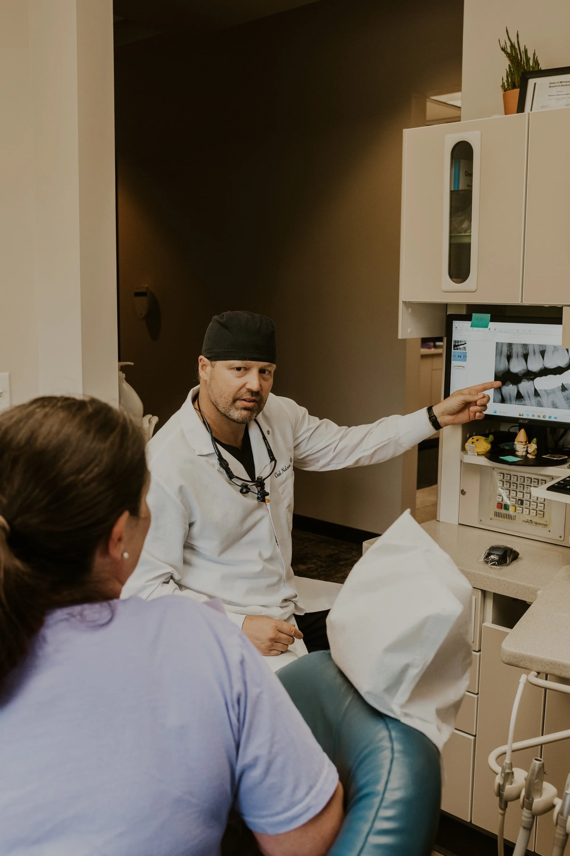 A dentist in a white coat shows dental X-ray images on a screen to a female patient seated in a dental chair.