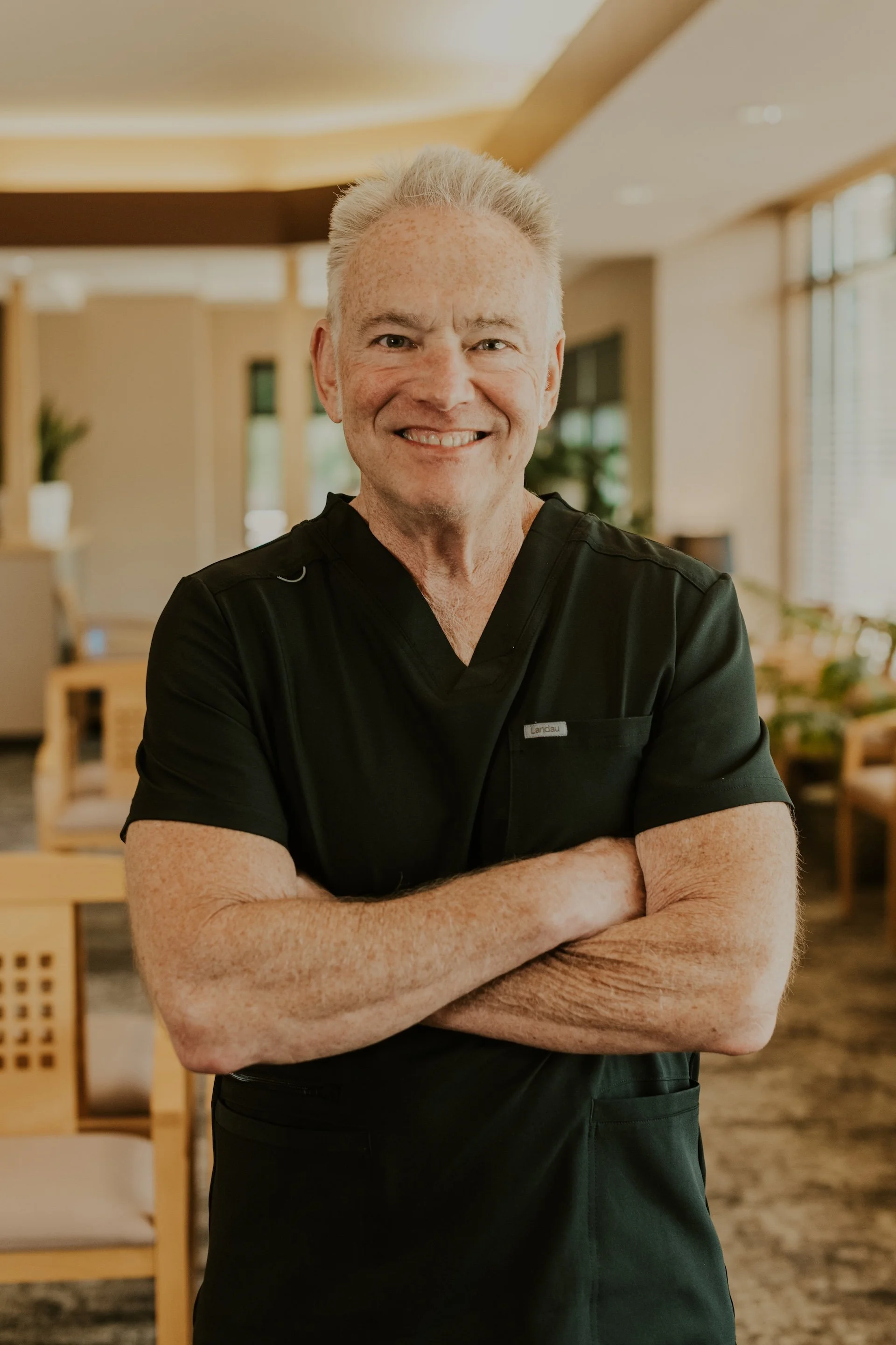 Dr. Gary Jenos smiling while wearing black scrubs and standing in the warmly lit Lakeville Family Dental office.
