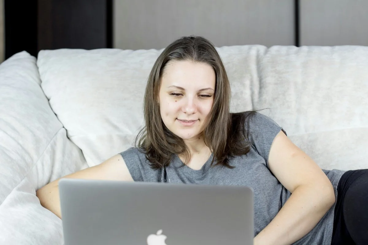 Young woman with brown hair, wearing a gray t-shirt, lying on a white couch, looking at a silver Apple laptop.