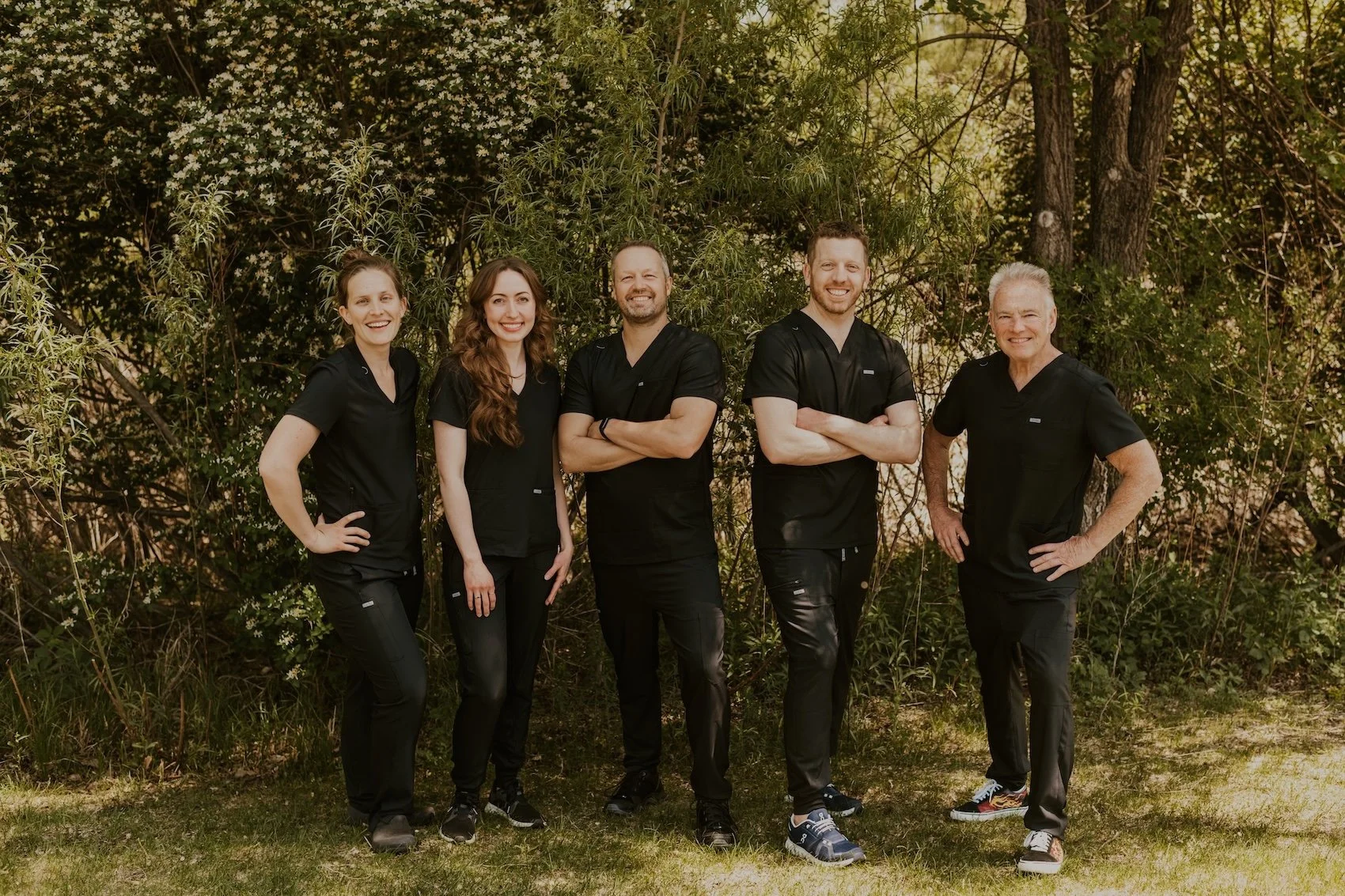 The Lakeville Family Dental team standing outdoors in front of trees, dressed in black scrubs, smiling at the camera.