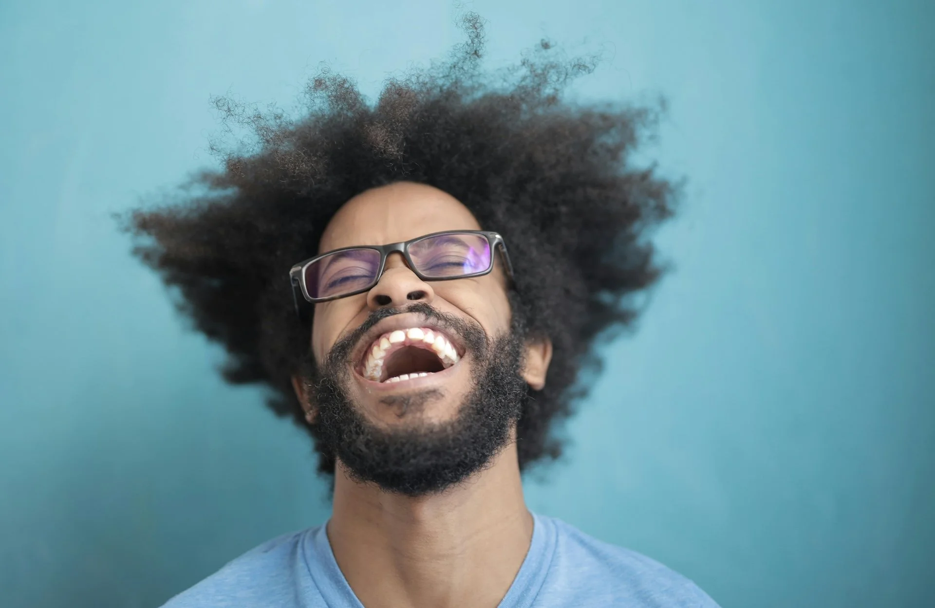 Man with curly hair and glasses, smiling broadly with mouth open, against a blue background.