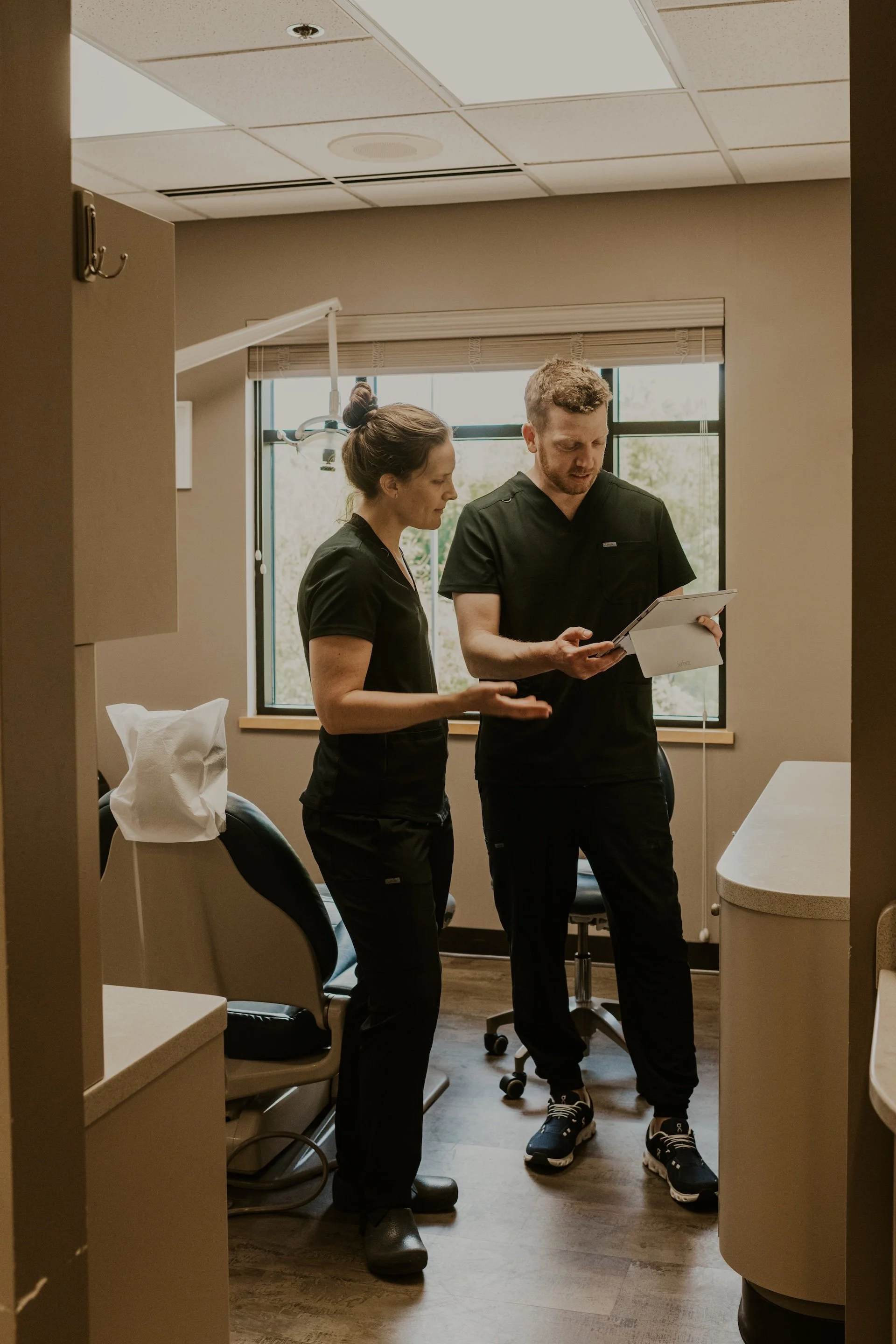 Two dentists standing in a dental office, reviewing a clipboard, with dental equipment and a window in the background.