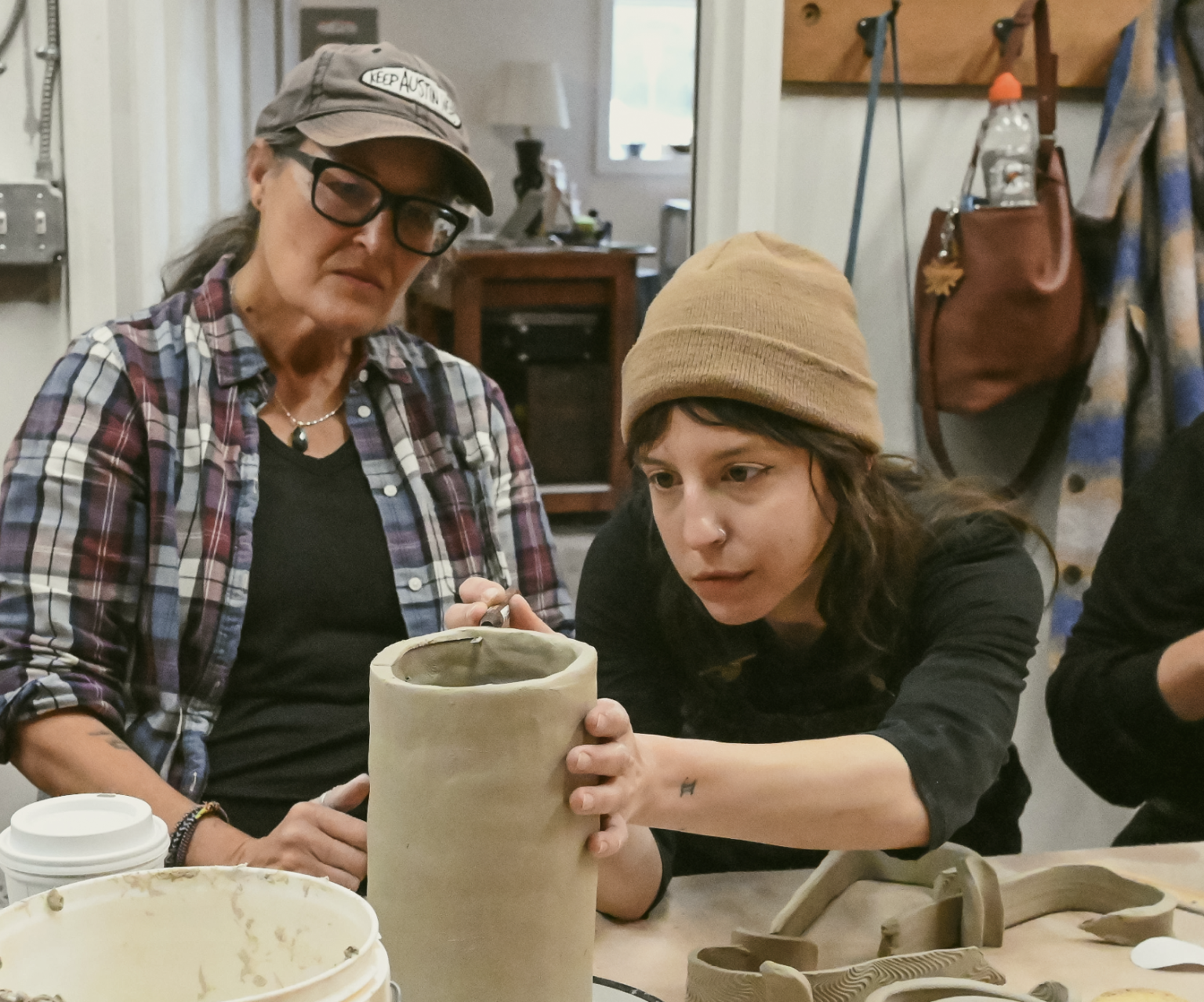 Two women working on a pottery project at a table, one holding a cylindrical clay piece, in a ceramics studio.
