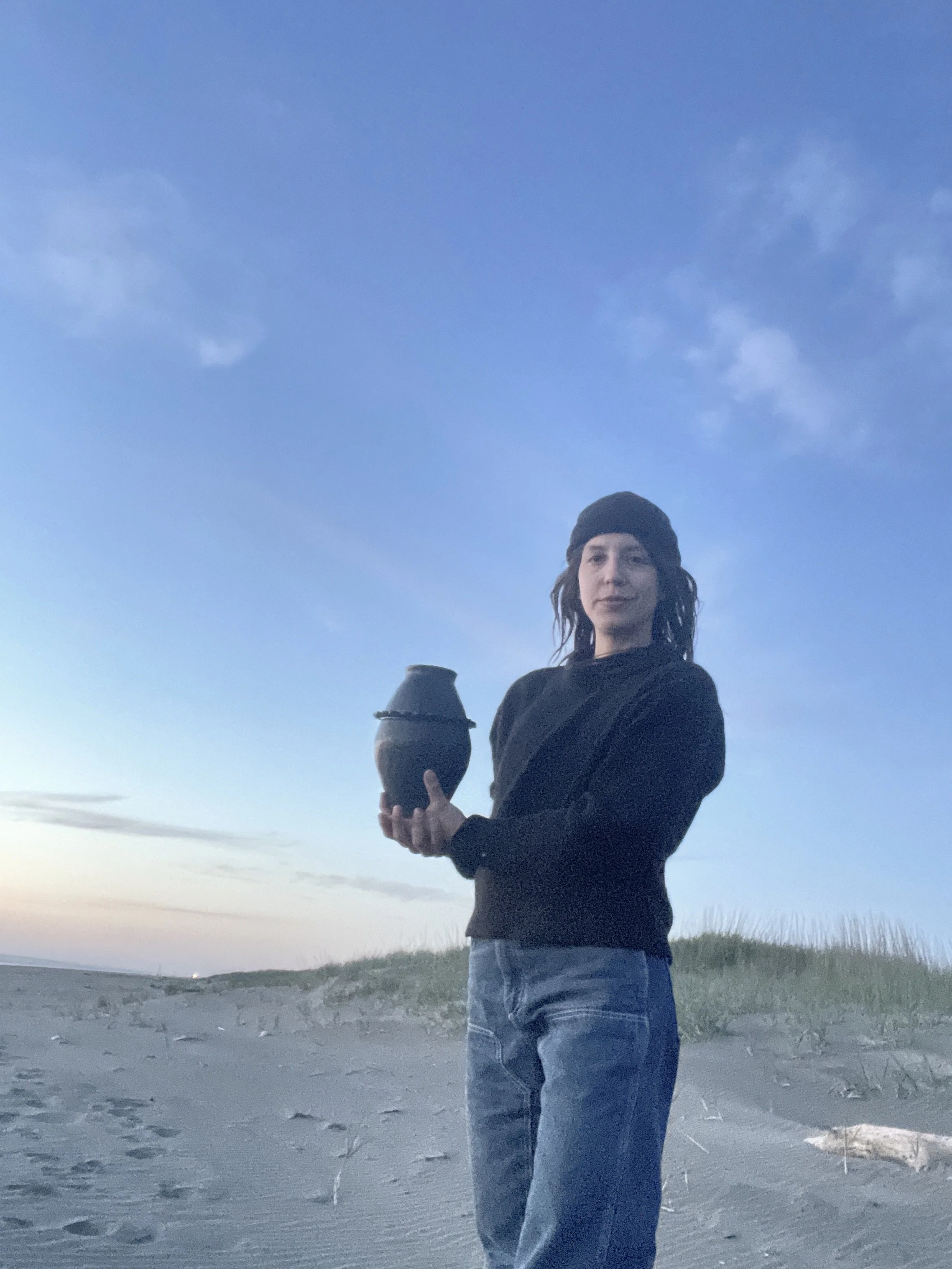 A woman wearing a black beanie and black sweater stands on a sandy beach holding a black ceramic vessel, with a late evening or early morning sky in the background.