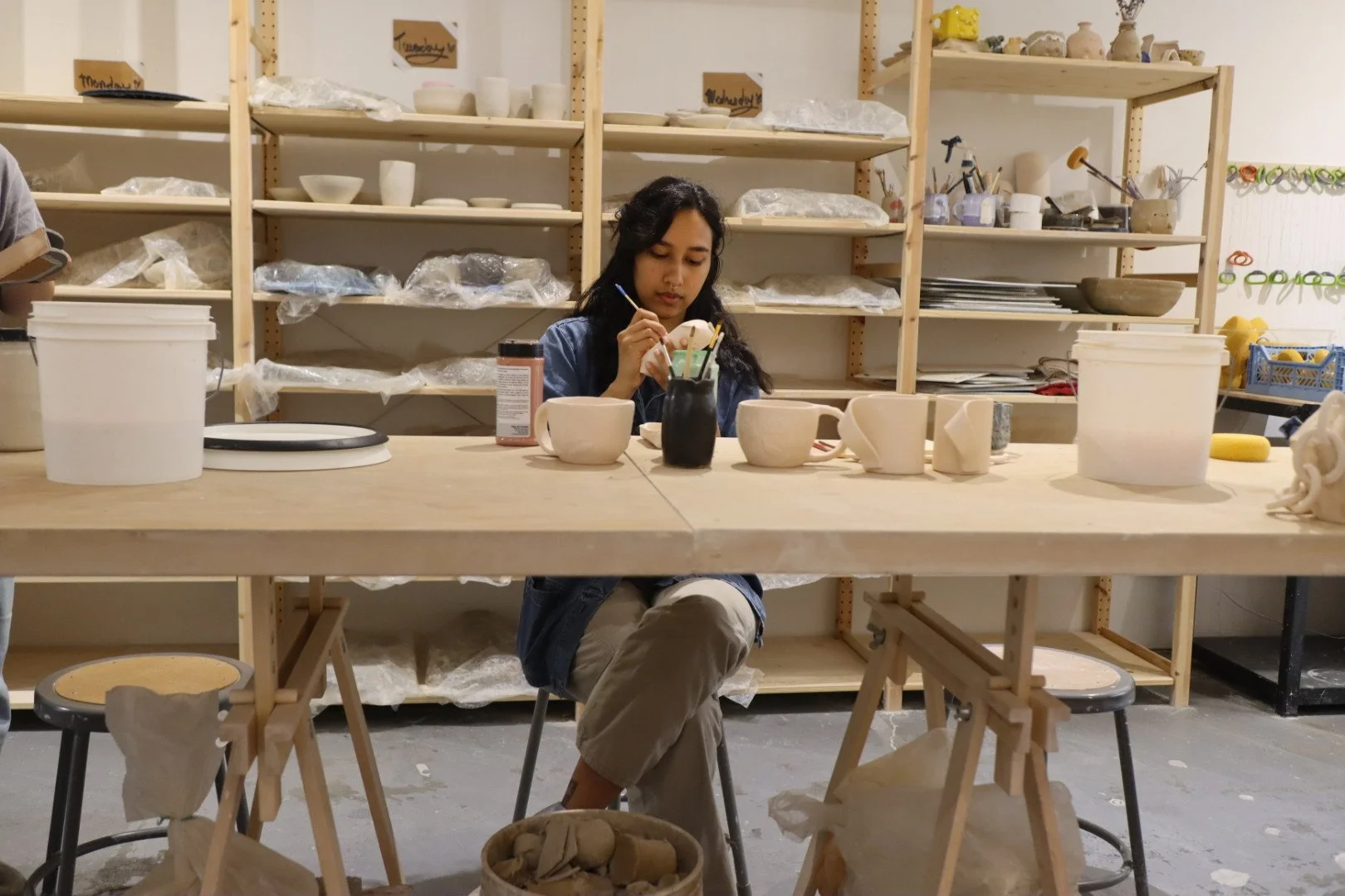 A woman sitting at a wooden work table in a ceramics studio, painting a ceramic piece surrounded by various unpainted ceramic cups, buckets, and tools, with shelves filled with pottery supplies in the background.