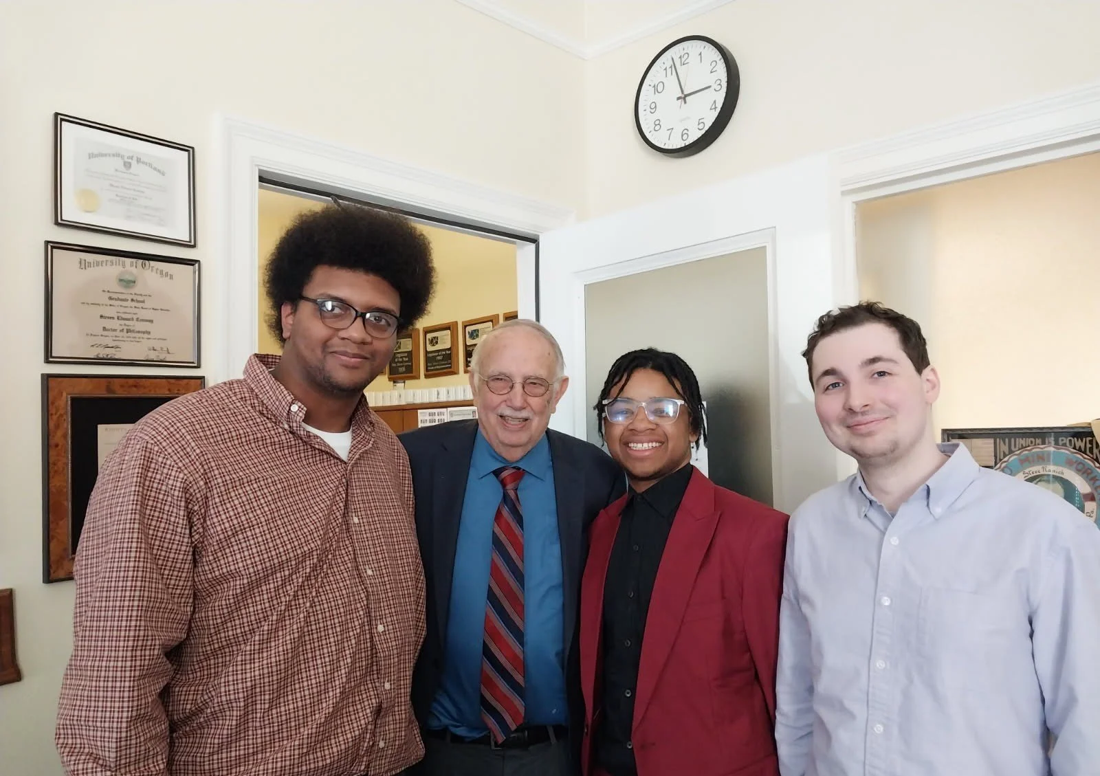 Four people standing together indoors, smiling at the camera, with framed certificates on the wall behind them.