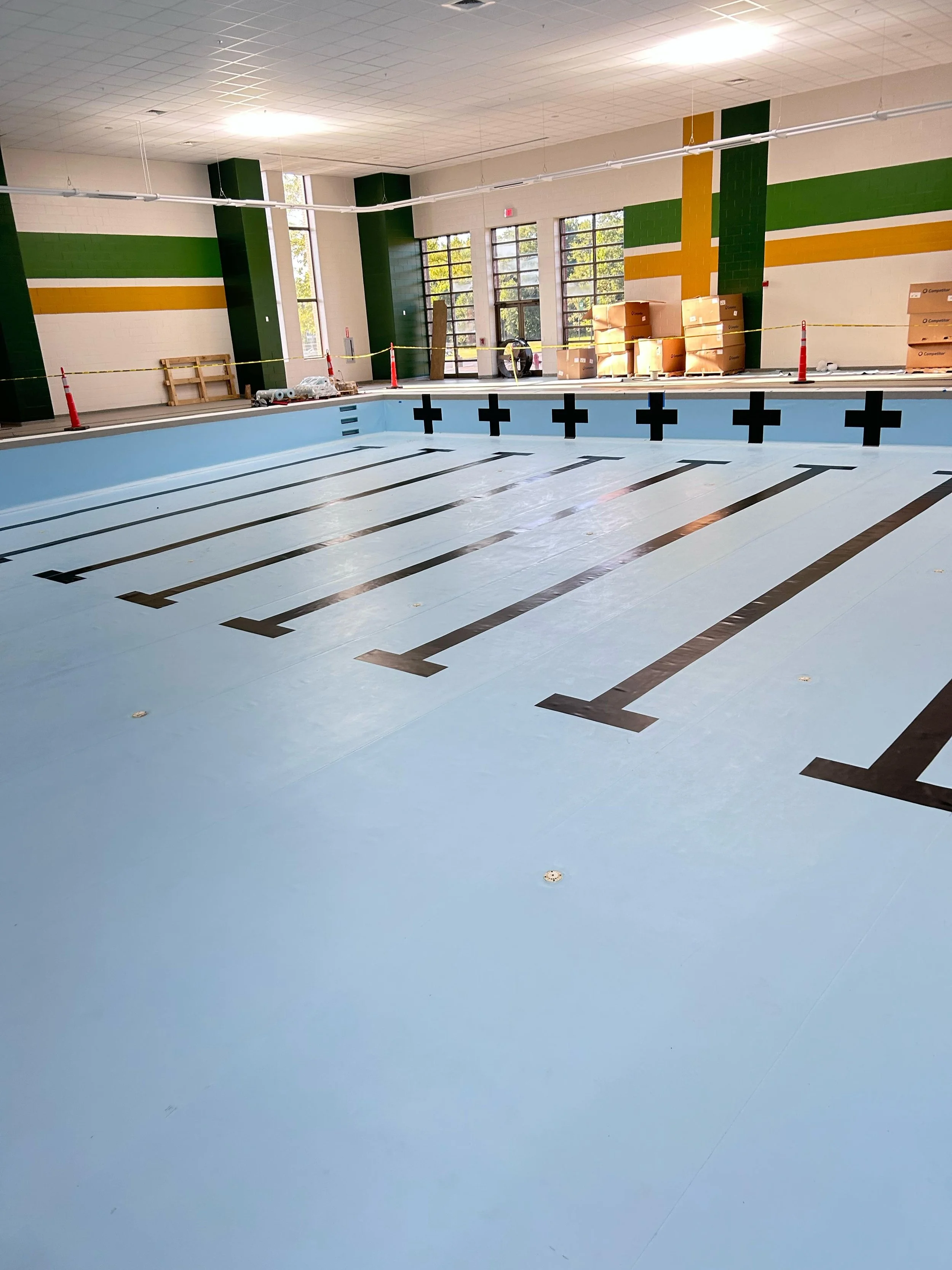 Empty indoor swimming pool under construction with lane markings and safety cones, large windows letting in natural light, and construction materials and boxes on the side.