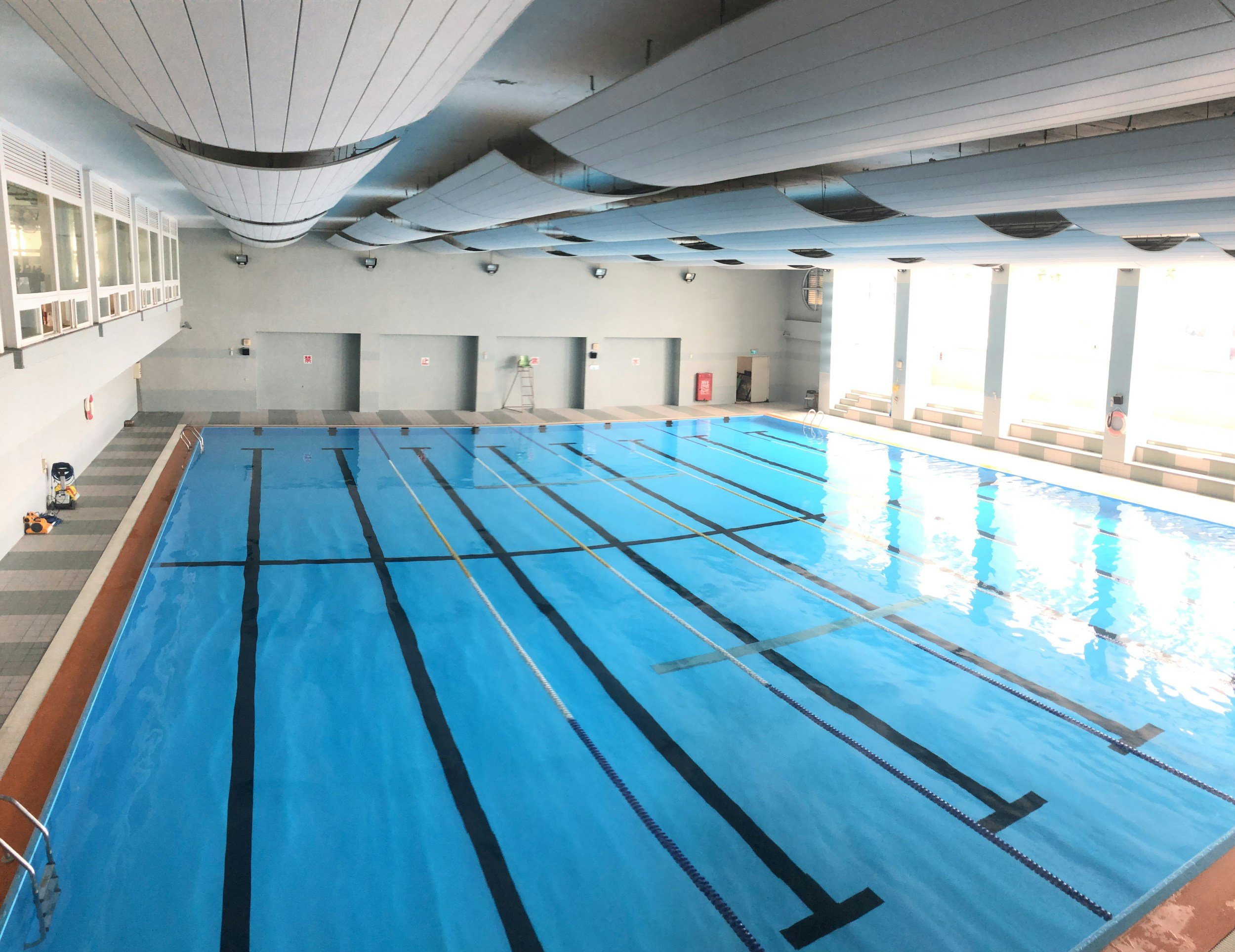 Indoor swimming pool with lanes, covered with a blue lining, light coming through large windows, with ceiling panels and safety equipment along the walls.