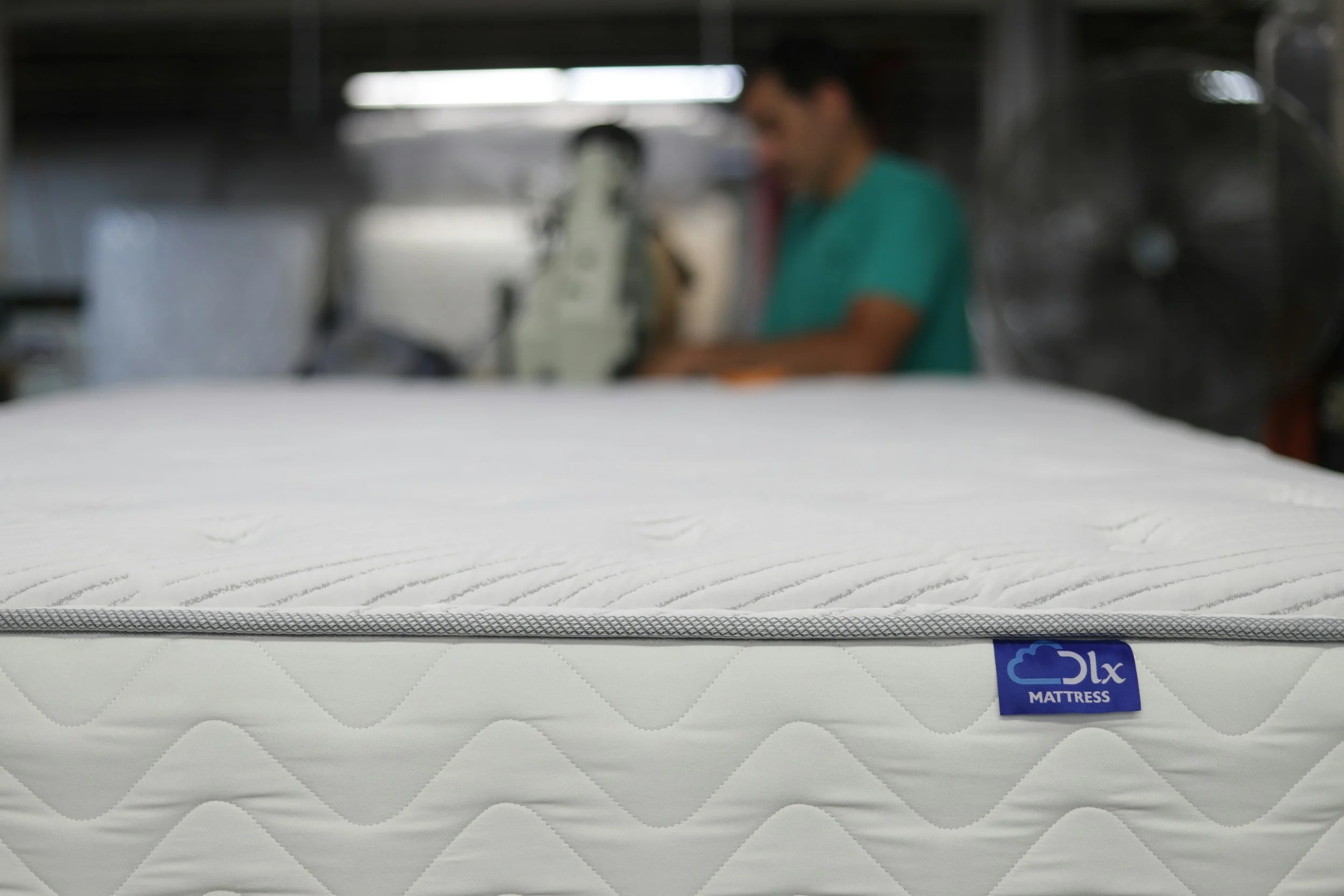 Close-up of a white mattress with a blue "OLX Mattress" label, with a blurred background of a person working with machinery.