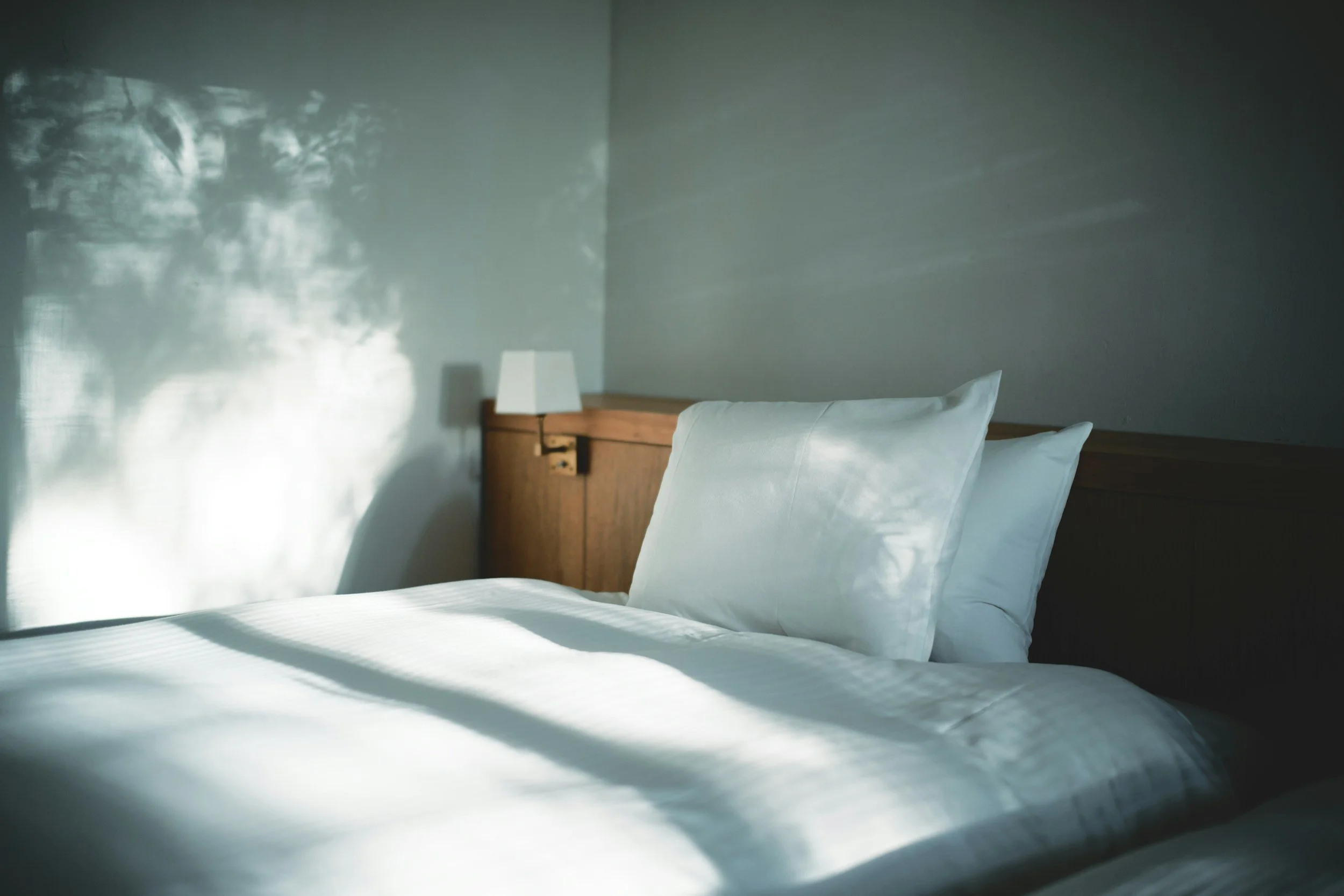 A neatly made bed with white linens and pillows in a minimalist bedroom, illuminated by natural sunlight casting shadows on the bed and wall, with a wooden headboard and a small white lampshade on a bedside table.